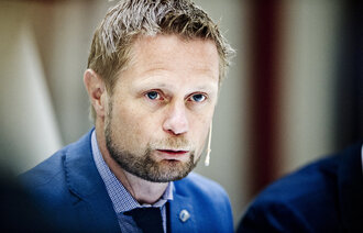 Portrett av Bent Høie med kikrofon og blå dress, lener seg mot bordet 