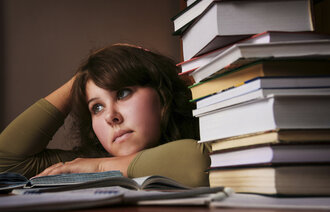 Bildet viser en student med et litt oppgitt uttrykk, som ligger over en oppslått bok på pulten. Det ligger en stabel bøker ved siden av henne.