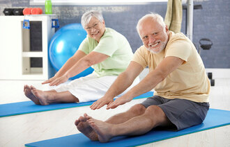 Bildet viser to eldre mennesker, en mann og en dame, som sitter på matter og tøyer ut.