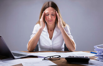 Bildet viser en kvinne som sitter foran et skrivebord med forskjellige permer og rekvisita på og holder seg til tinningene