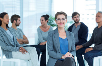 Bildet viser en gruppe som sitter i en halvsirkel og en kvinne som sitter med ryggen til gruppen og ser på oss