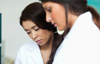 Bildet viser to kvinnelige sykepleiere som har ikke-etnisk norsk opprinnelse, som står og ser på noe sammen.