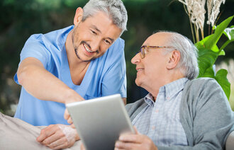 Sykepleier og eldre mann smiler og ser på ipad.