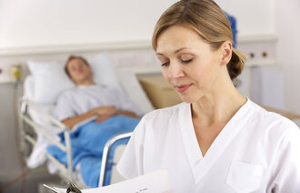 Bildet viser en sykepleier som står foran en seng på sykehuset med en pasient i.