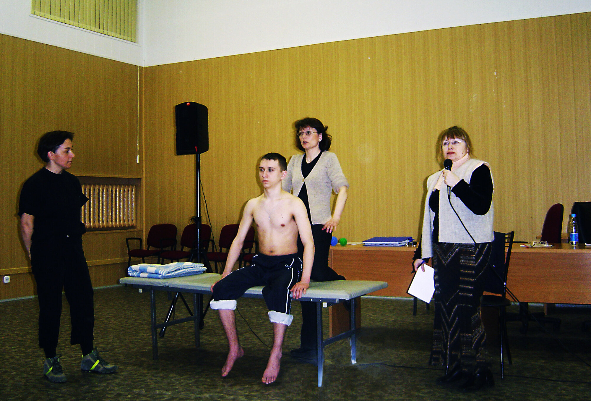 Bildet viser to norske fysioterapeuter som demonstrerer foran et russisk publikum. En mannlig pasient sitter på en seng, mens en tolk oversetter.