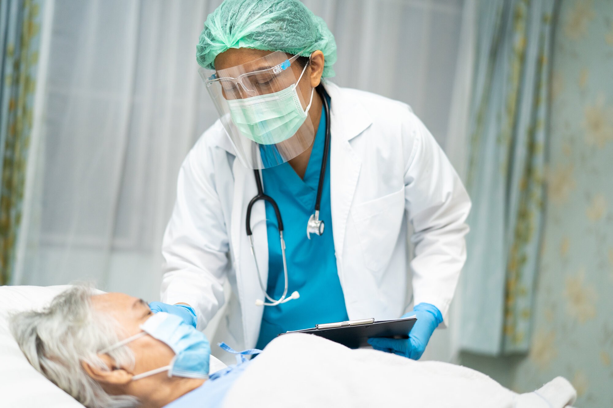 Bildet viser en eldre mann som ligger i senga med maske på, mens legen står ved siden av med maske og visir