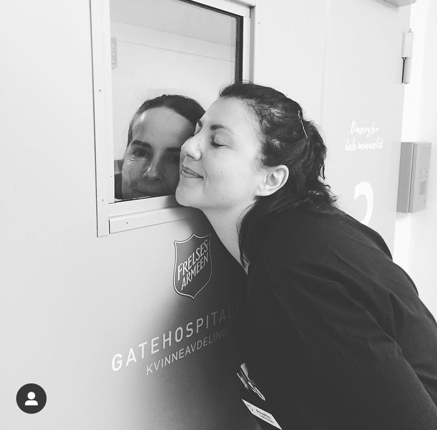 Bildet viser to sykepleiere som gir hverandre en klem gjennom en dør.