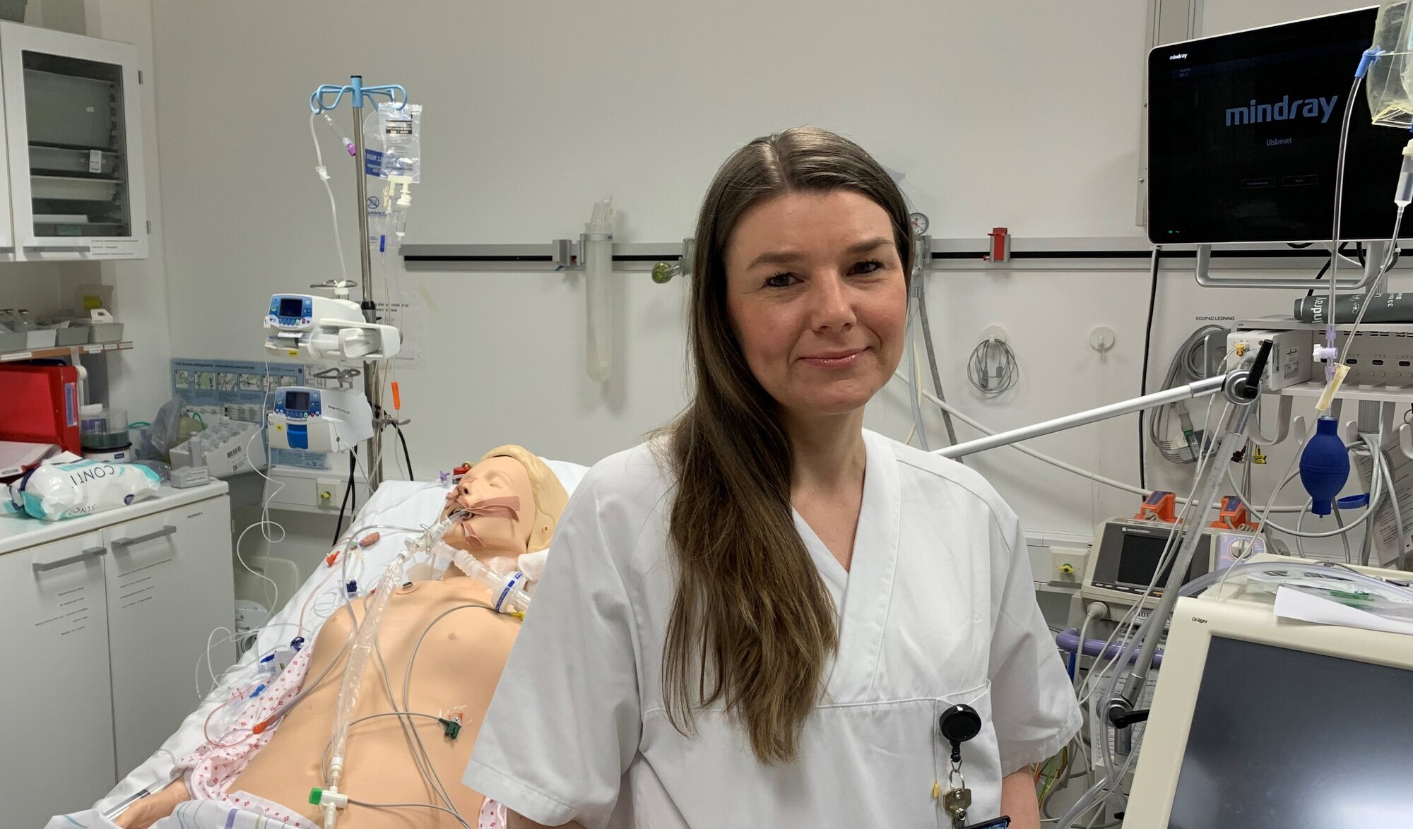 Bildet viser Monica Thallinger foran en sykehusseng, der det ligger en simuleringsdukke.