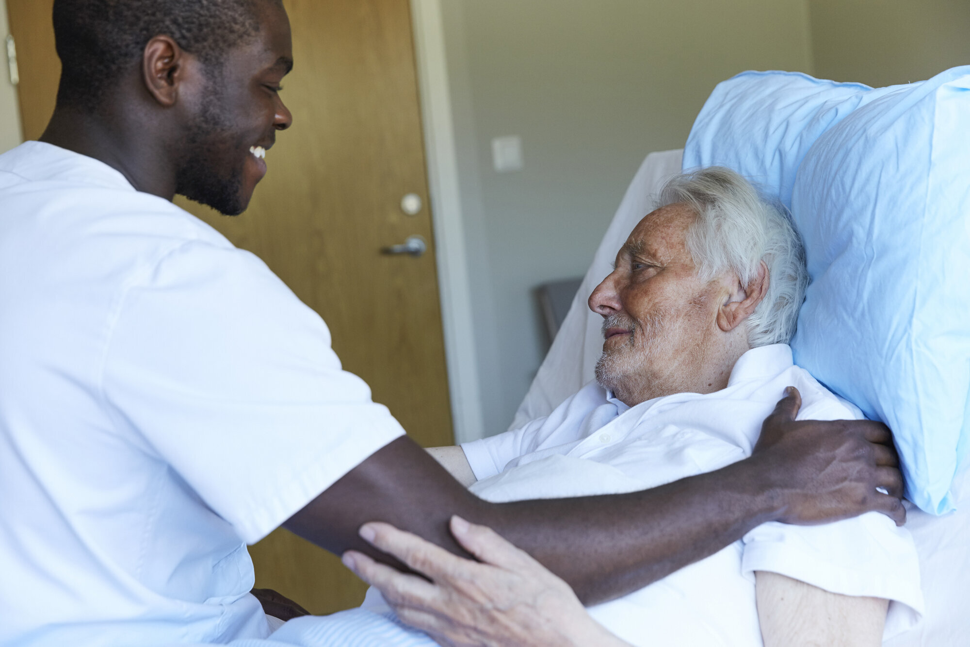 Bildet viser en mannlig sykepleier som smiler og holder hånden sin på skulderen til en liggende pasient