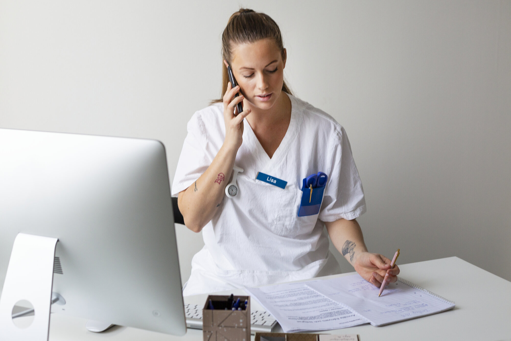 Bildet viser en ung, kvinnelig sykepleier som snakker i telefonen og tar notater.