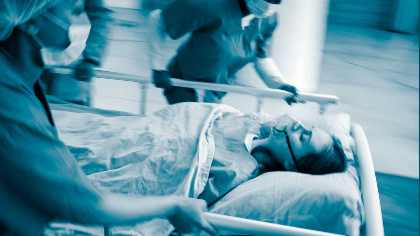 The photo shows a patient on a stretcher carried by running health professionals. The patient is wearing an oxygen mask.