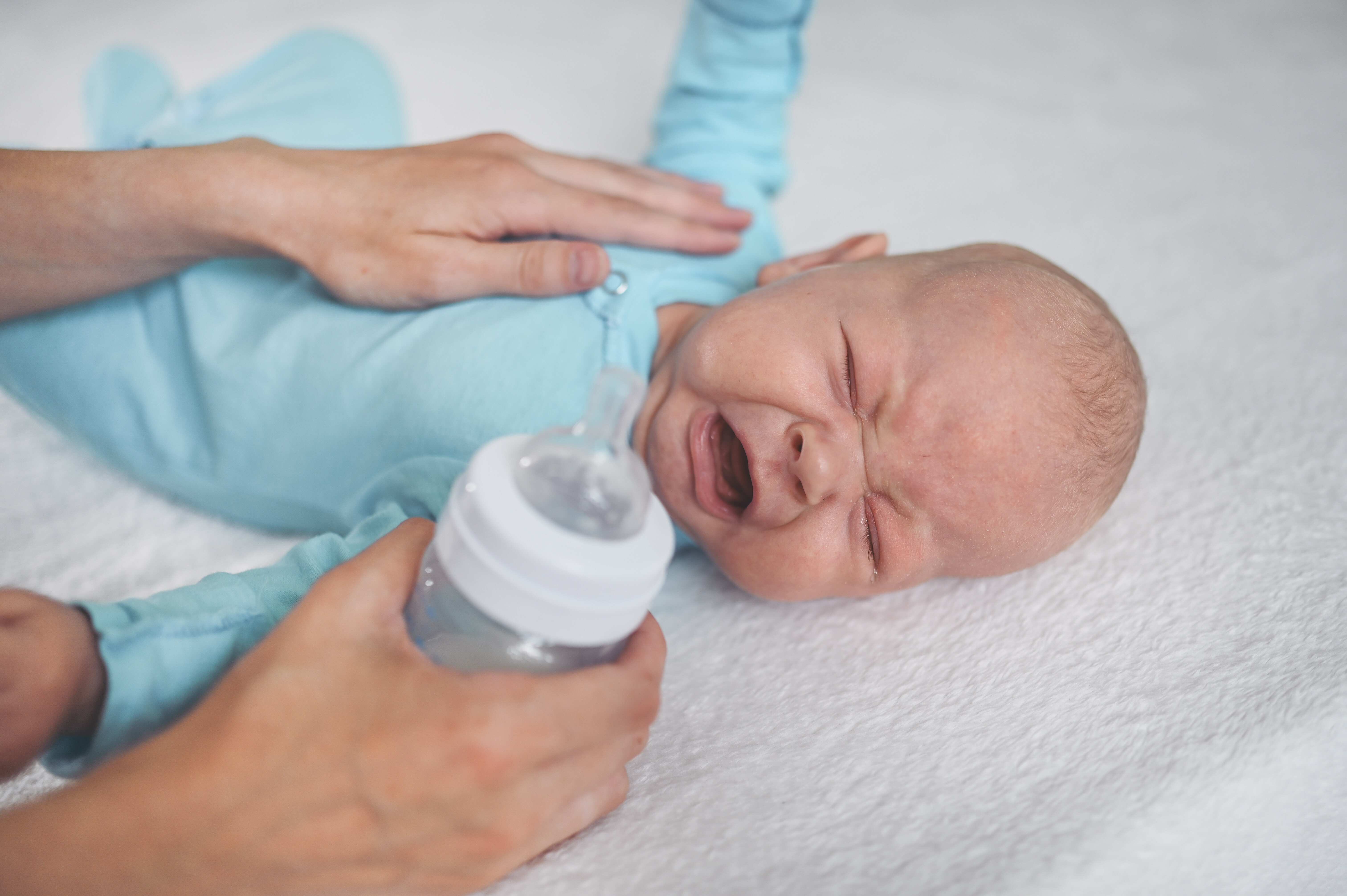 The photo shows a crying infant. A soothing hand lies comforting on the baby's stomach. The other hand is holding a bottle ready to feed the baby