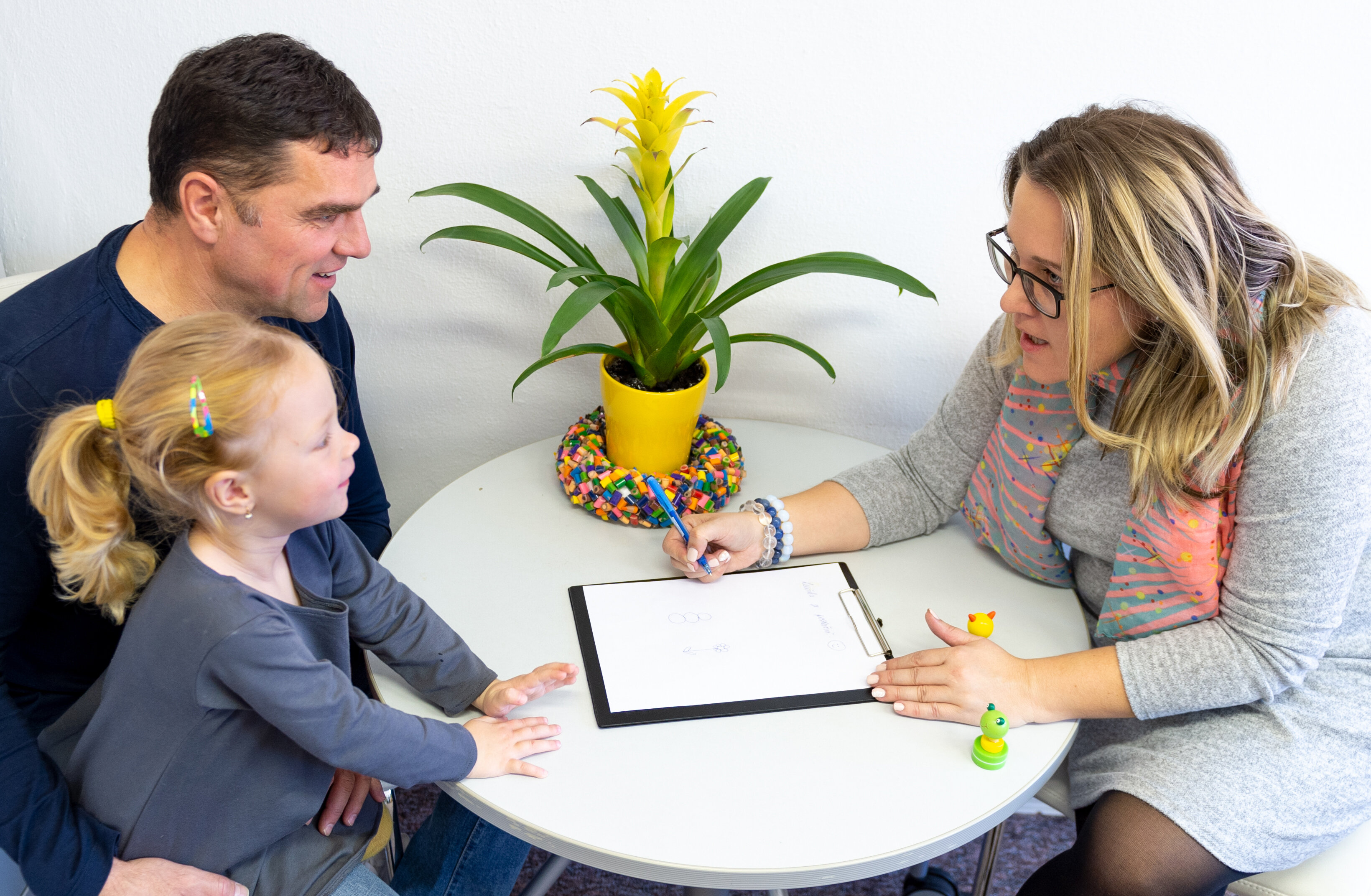 The photo shows a father with his daughter on his lap.  They are sitting at a table opposite a woman leaning towards them. She has a notebook in front of her.