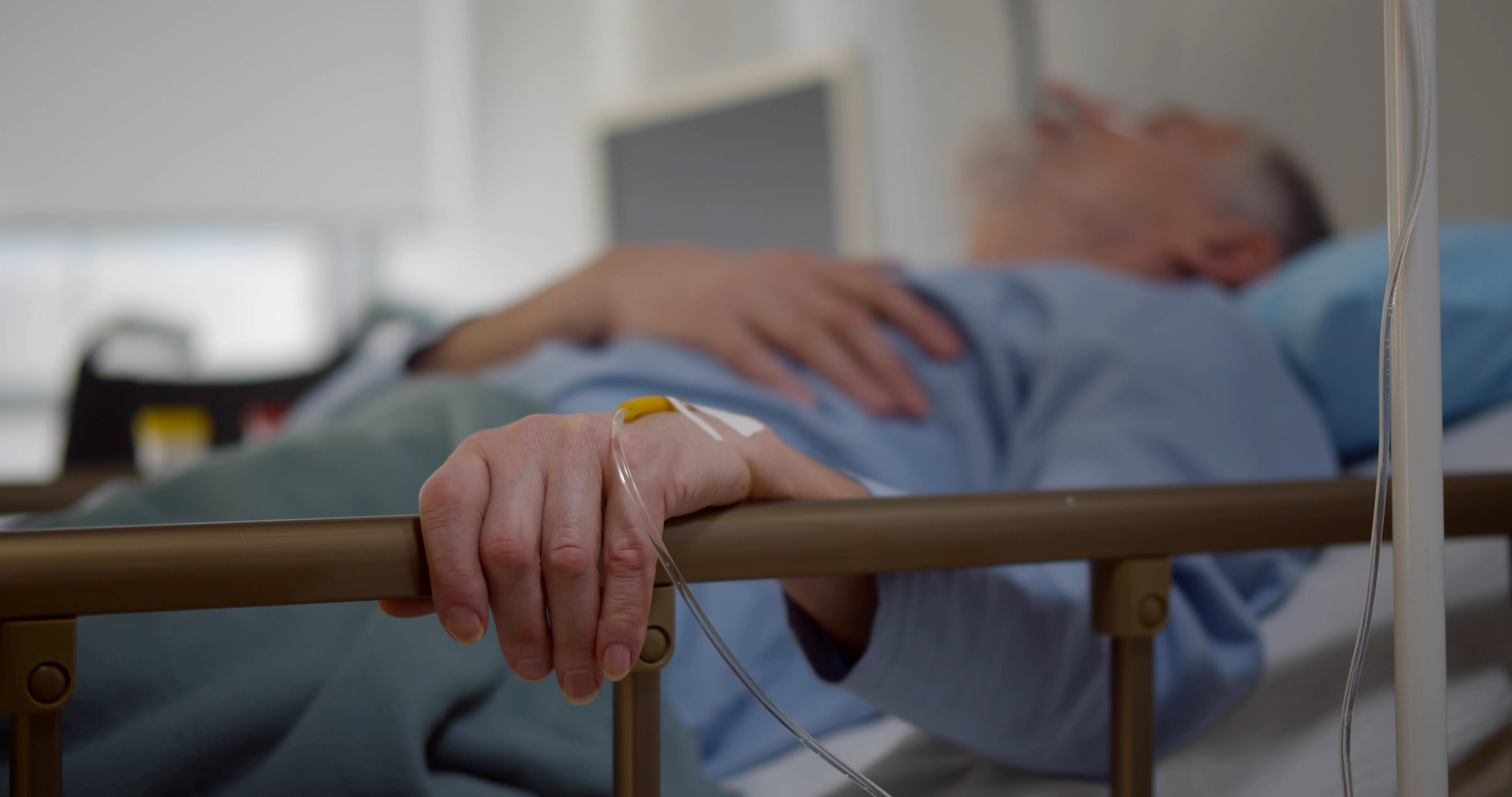 The photo shows an older man lying in a hospital bed. His left hand is resting on the bed rail. In his hand is a venous access port.