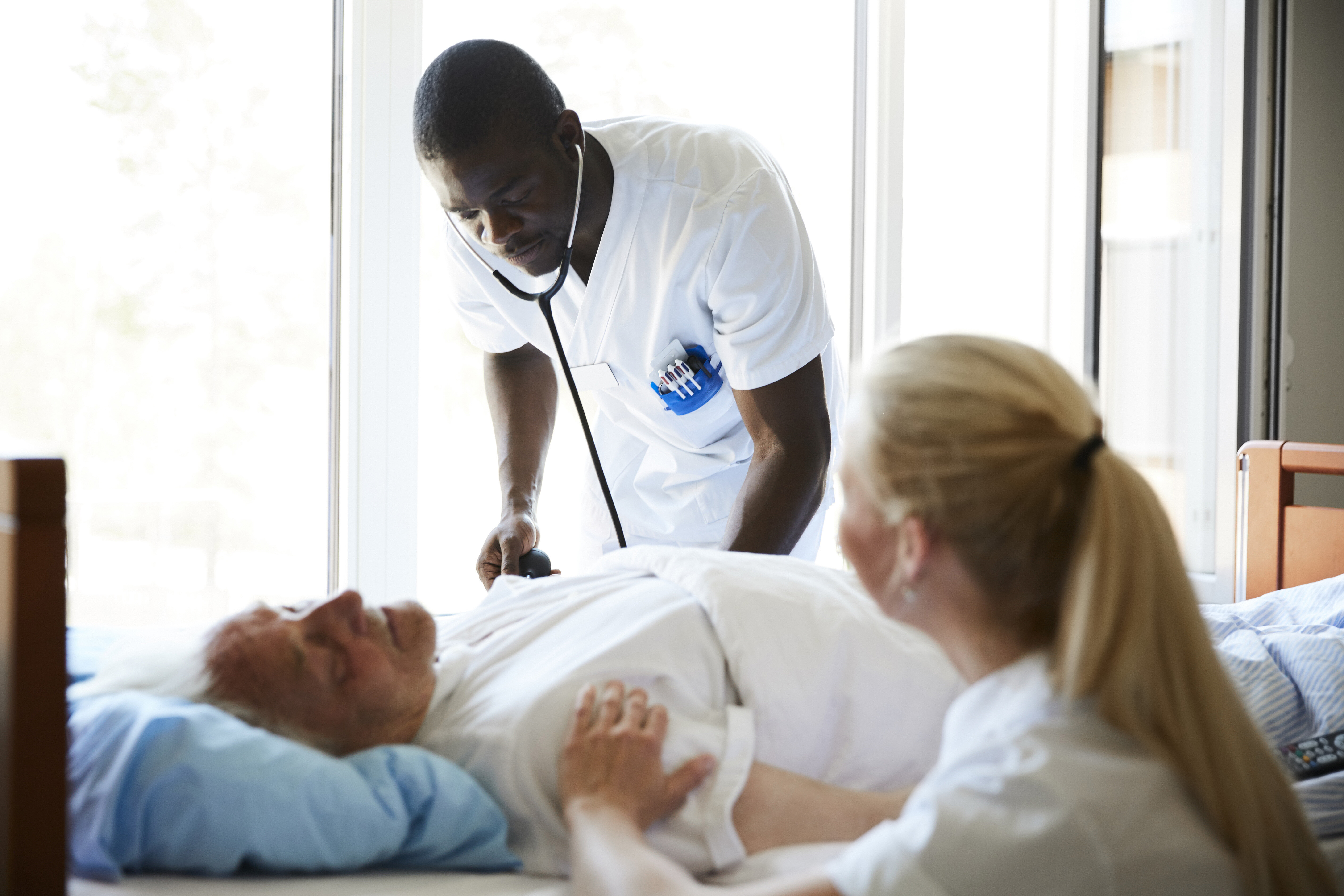 The photo shows an older patient lying in a hospital bed. A male nurse is bending towards him listening with a stethoscope. A female nurse is sitting beside the bed.