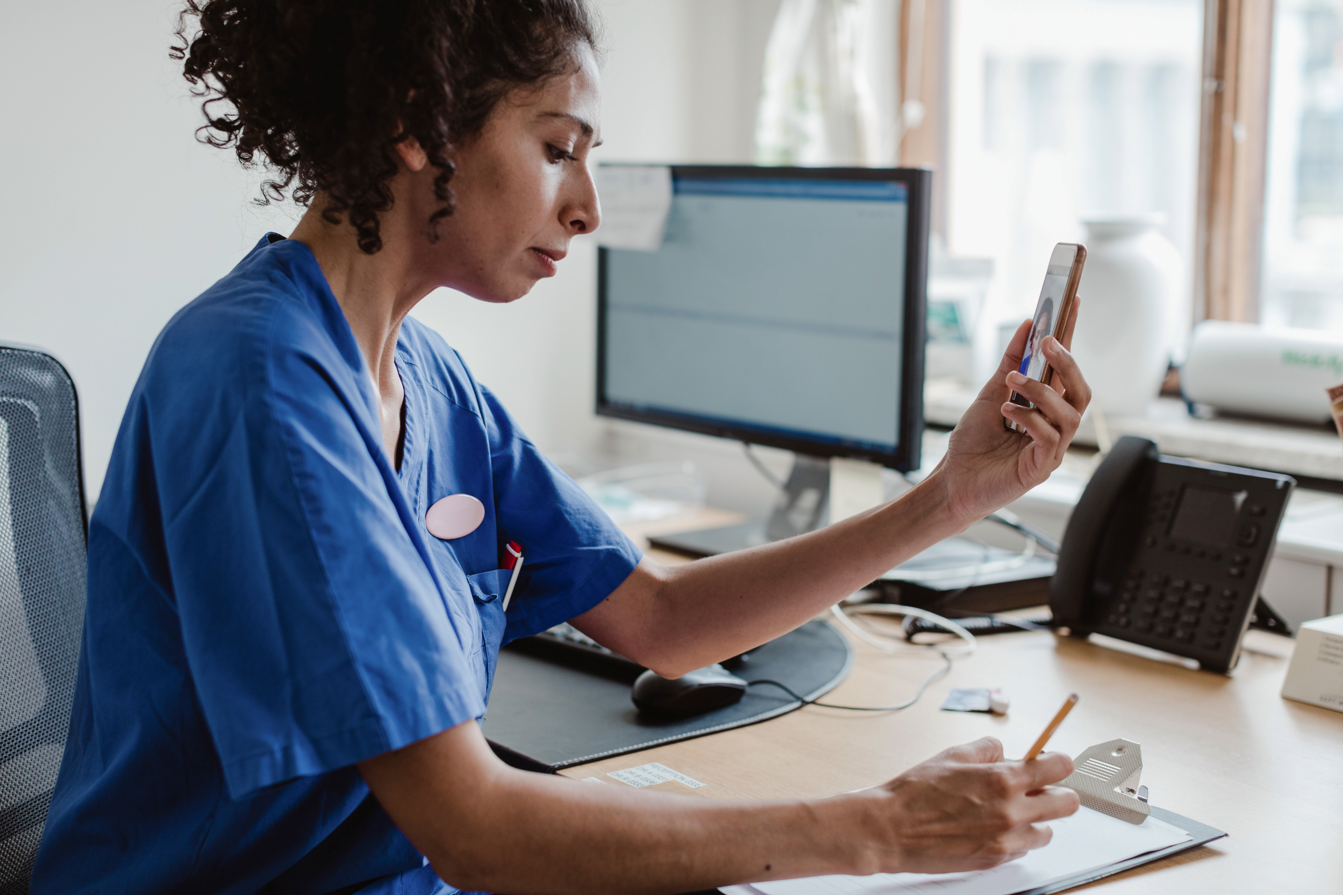 The photo shows a nurse having a video chat on her mobile phone. She is sitting at her desk writing a note.