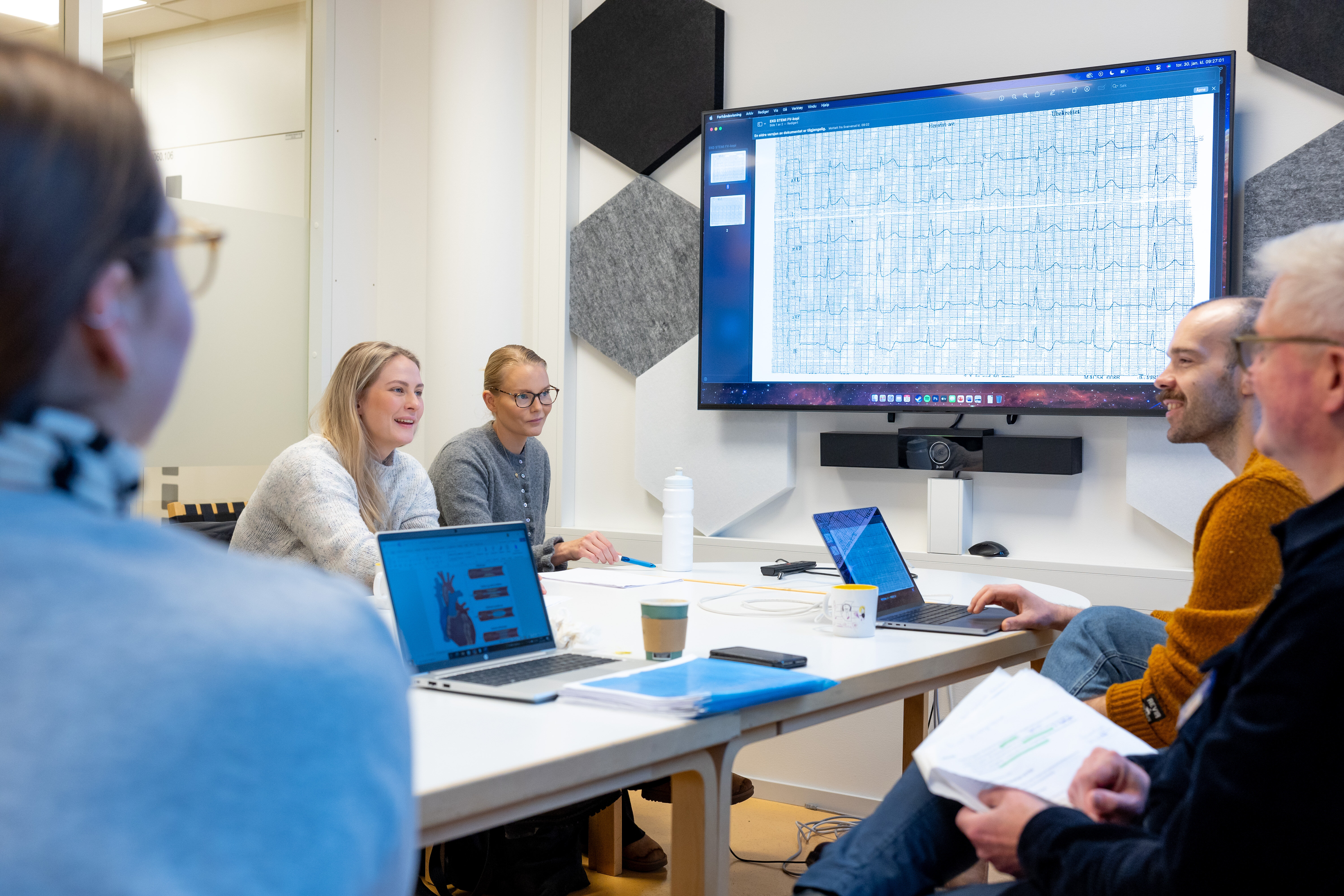 The photo shows two students sitting around a table opposite two academic supervisors. A third student can be seen with her back towards the camera. At the screen on the wall graphs of cardiac arrhythmias can be seen. The people are participating in a reflection group.