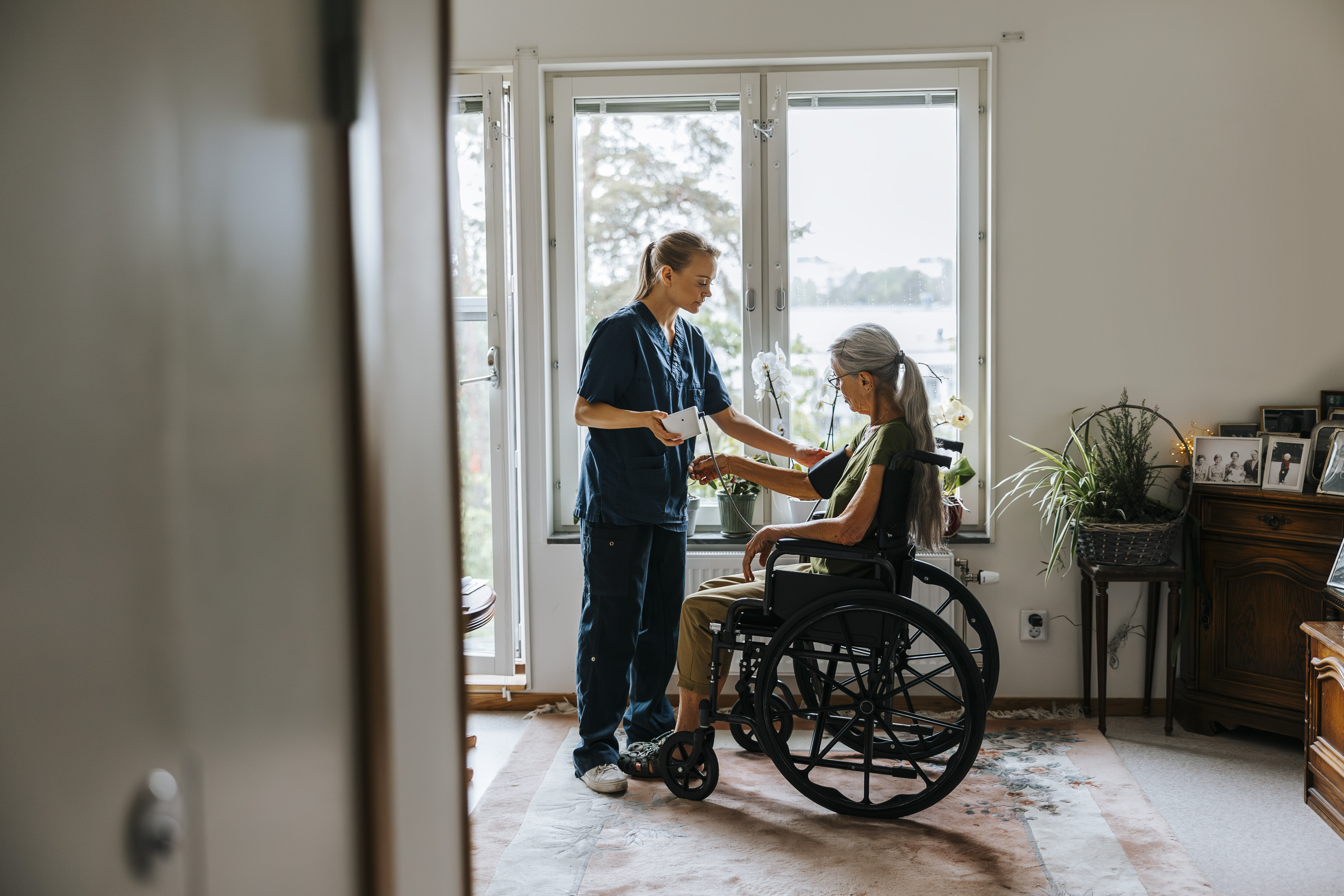 The phowo shows a nurse standing in front of a woman in a wheel chair. The nurse is monitoring her blood pressure. The are in front of a window in the patient's living room