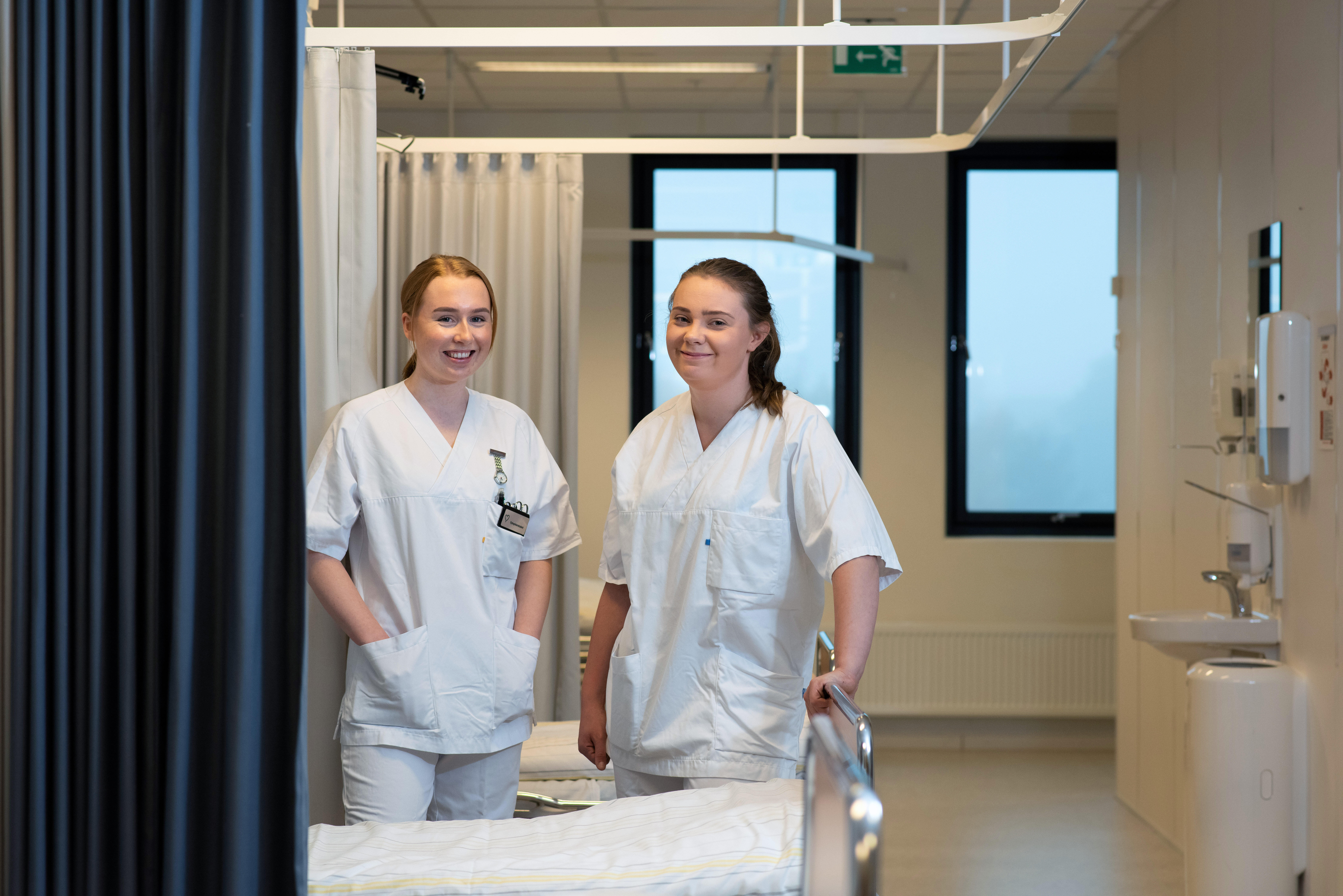 Bildet viser to sykepleiere på sengepost som ser mot kamera