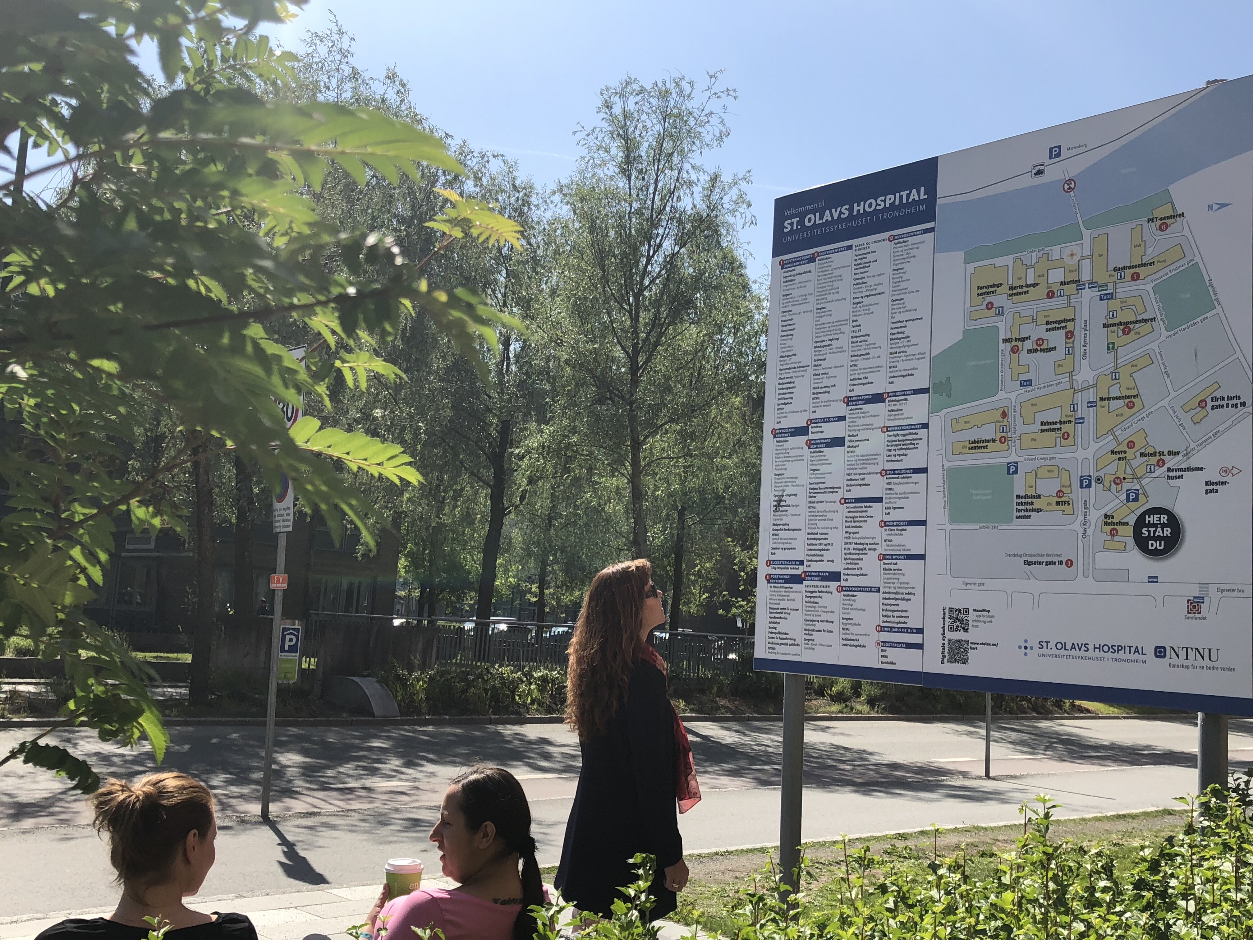 The picture shows a woman standing in front of a large sign outside St. Olavs Hospital. The sign displays information about the hospital.