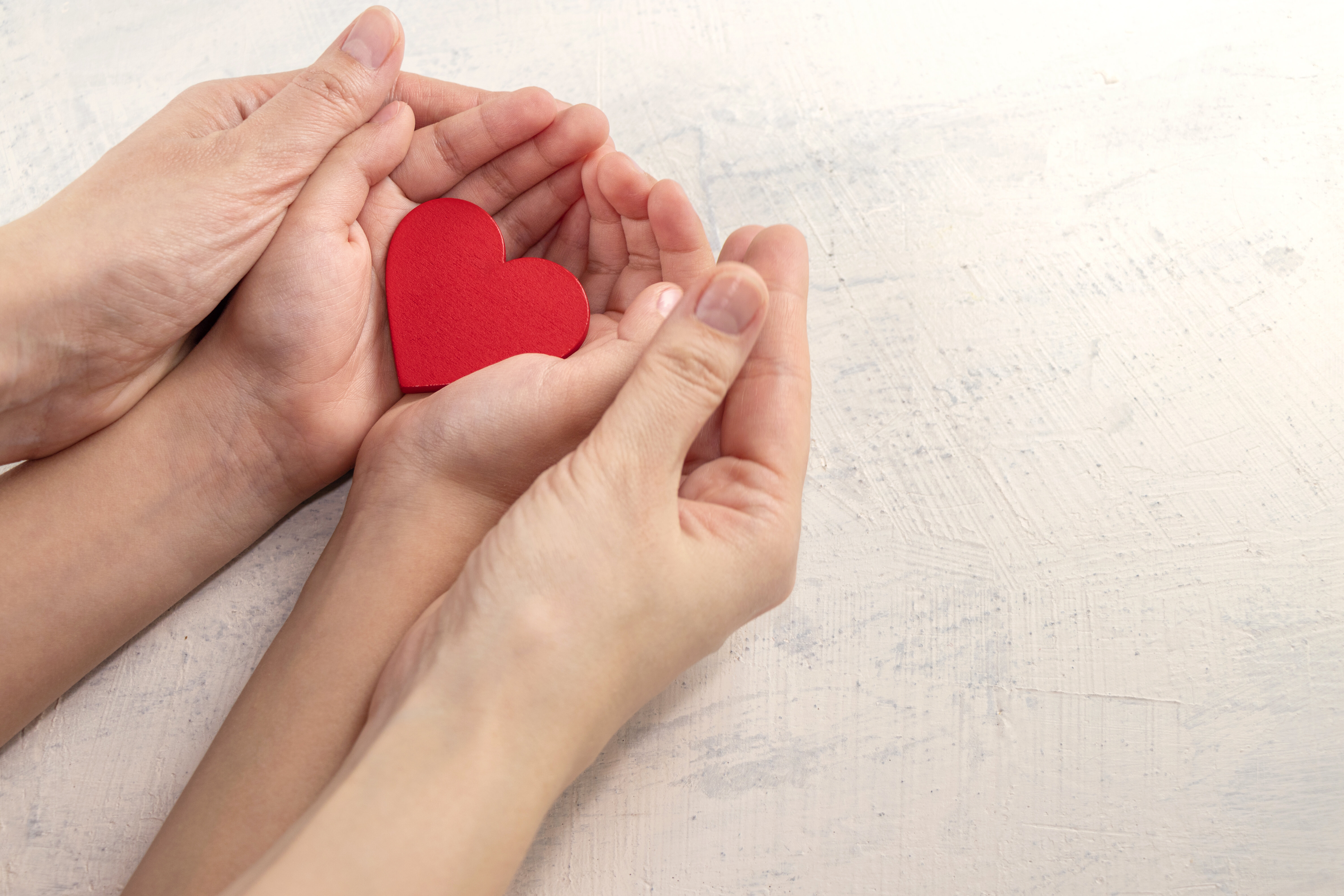The image shows a pair of female hands gently holding a child’s hands. The child is holding a heart in their hands