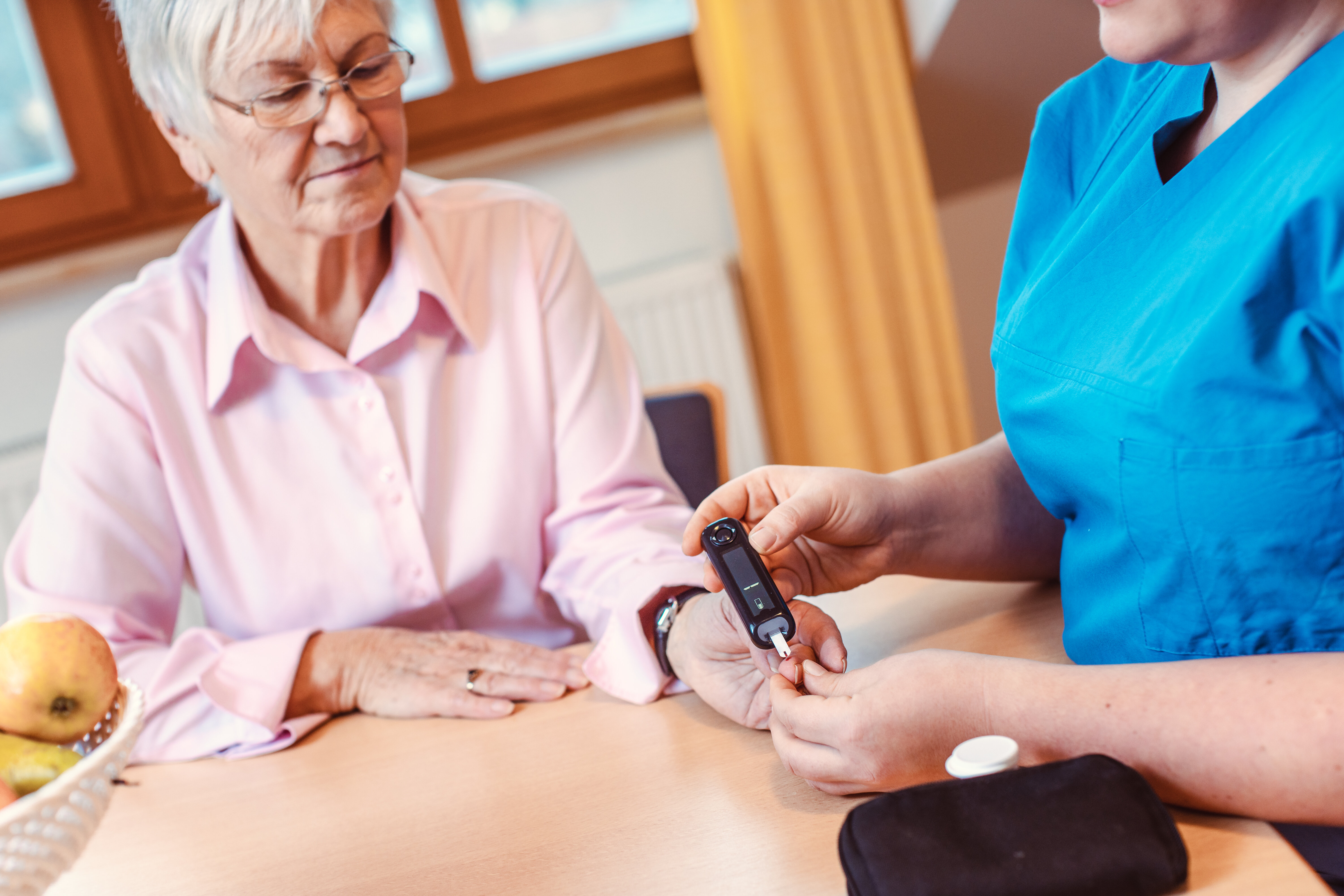 The image shows a nurse monitoring the blood sugar of an older woman in her home