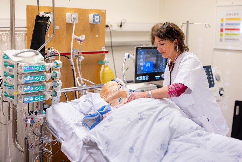 The image depicts a simulation mannequin connected to a ventilator. A nurse is leaning over the bed.