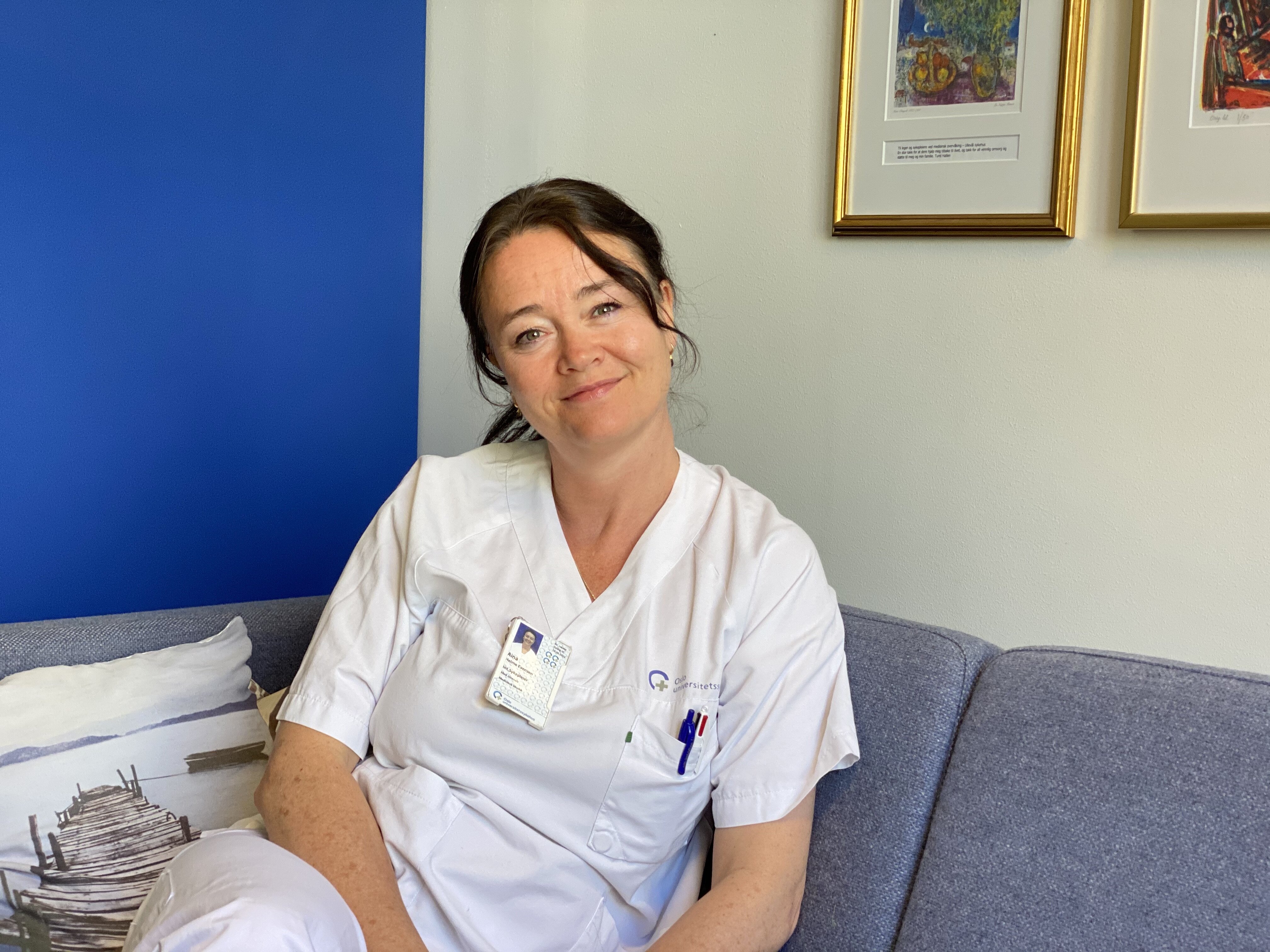 Bildet viser Aina Evensen Helme på jobb i sykepleieruniform. Hun sitter og ser inn i kameraet