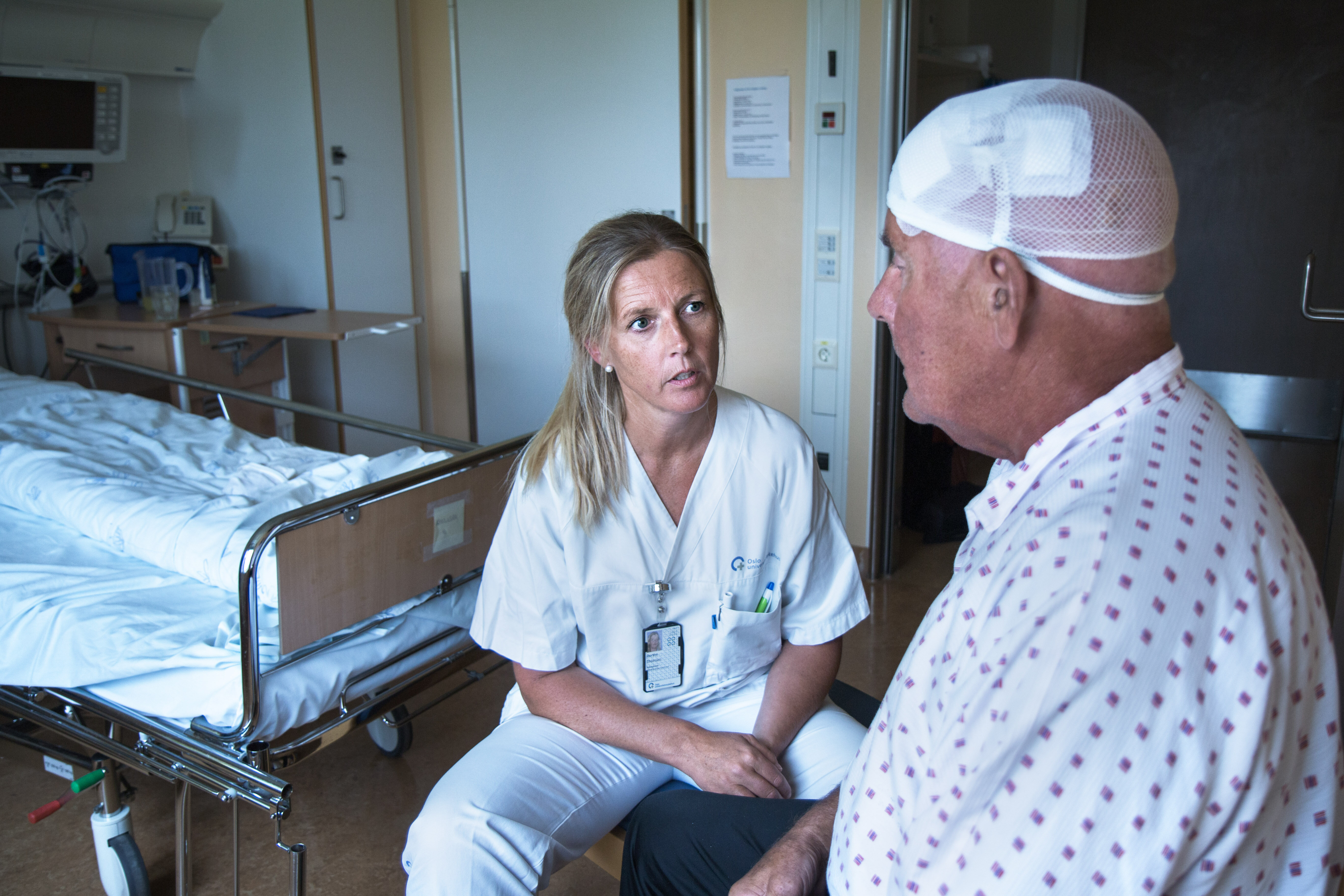 Bildet viser en sykepleier som sitter overfor en nyoperert parkinsonpasient. Han har bandasje rundt hodet