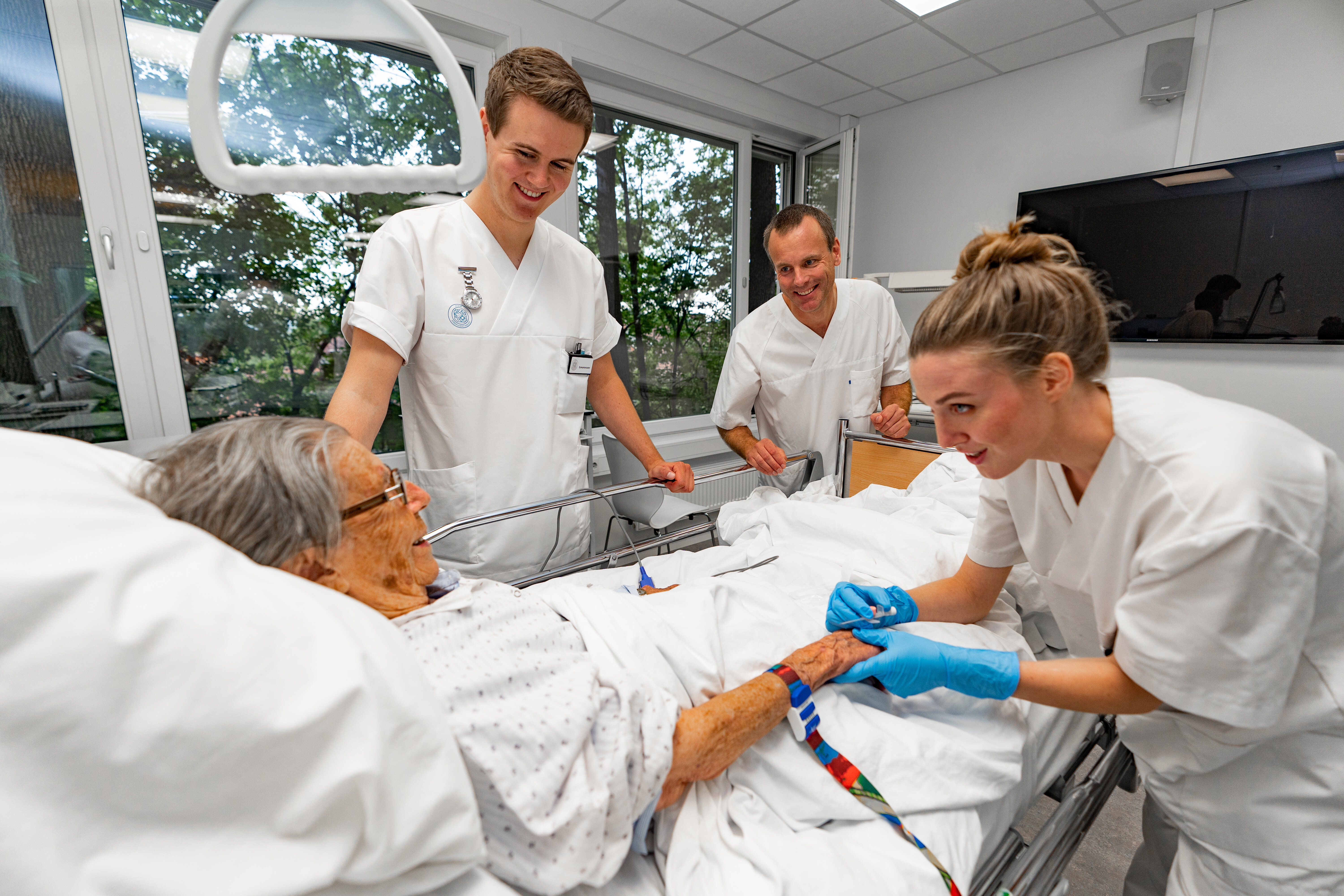 Bildet viser en eldre pasient som ligger i en sykehusseng. Rundt henne står to sykepleierstudenter og en veileder. Alle smiler, og den ene studenten holder pasientens hånd