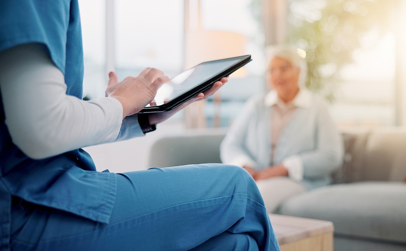 Bildet viser en sykepleier som sitter med en iPad med en pasient i bakgrunnen