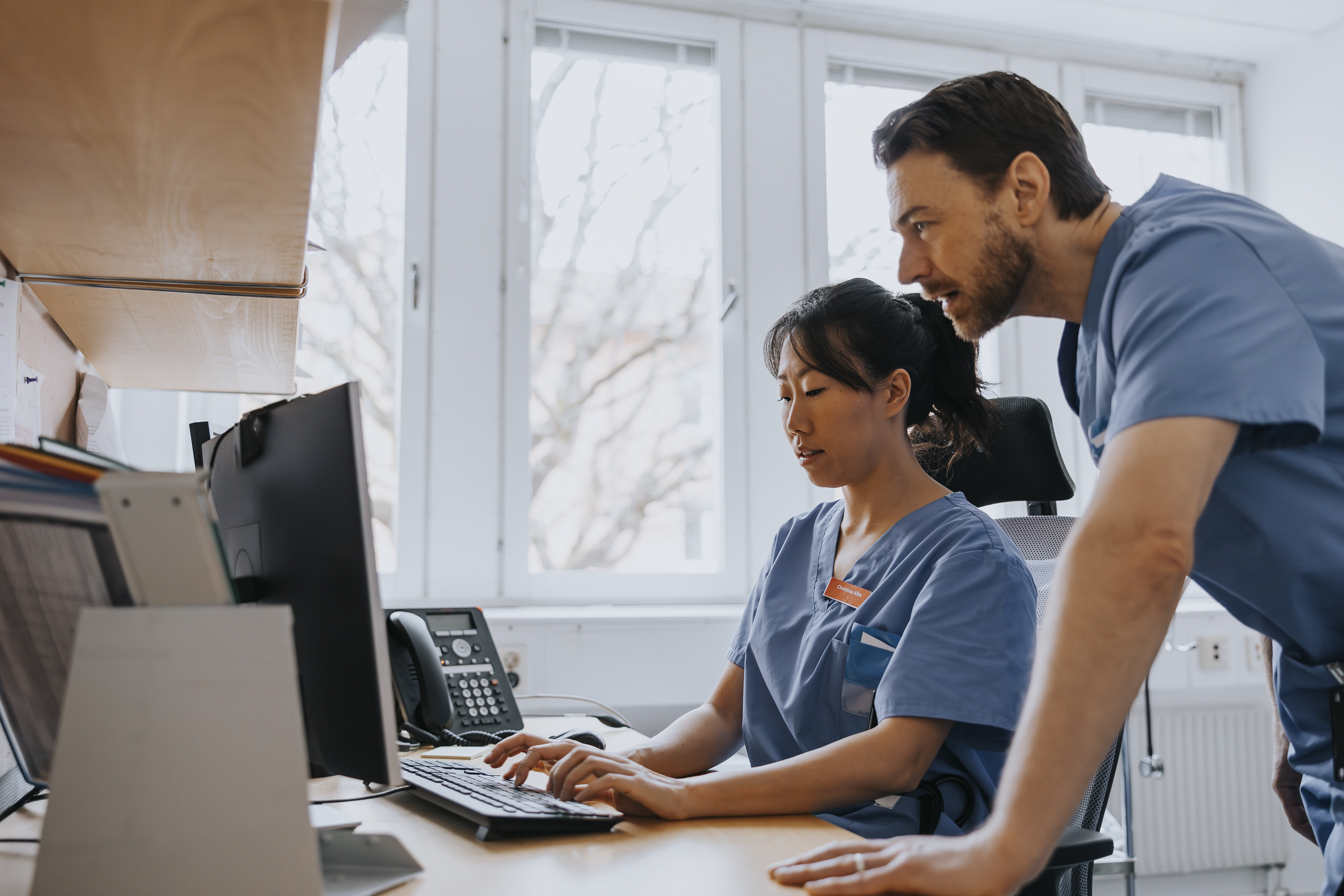 Bildet viser en kvinnelig sykepleier som sitter foran en PC. En mannlig sykepleier står bak henne og kikker på skjermen