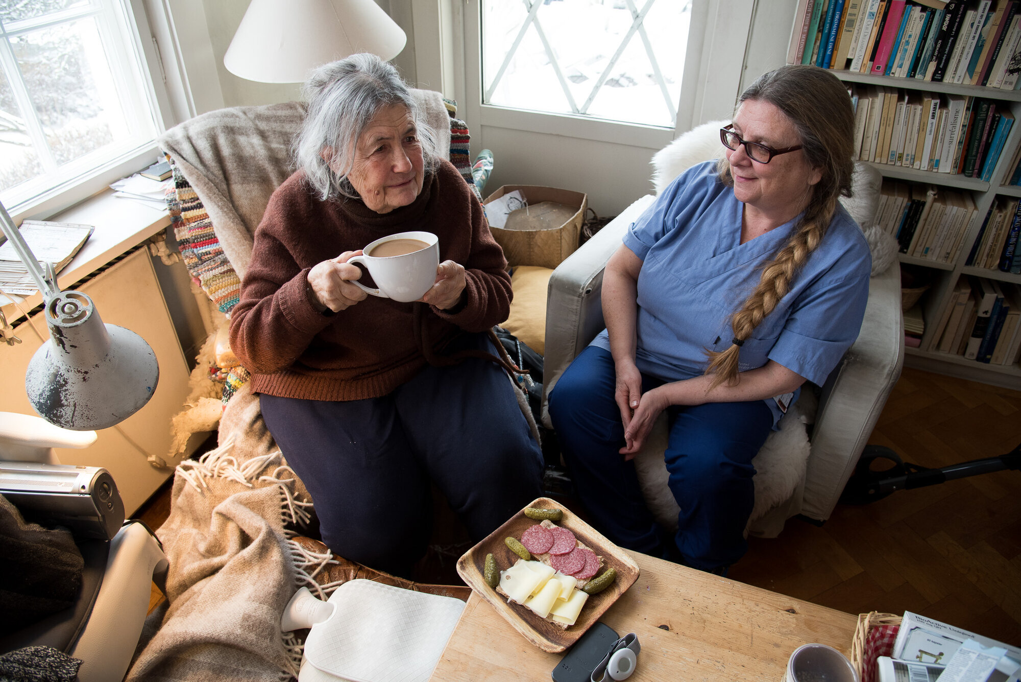 Bildet viser en beboer som drikker kaffe sammen med hjemmesykepleieren. Foran beboeren står et fat med påsmurte brødskiver.
