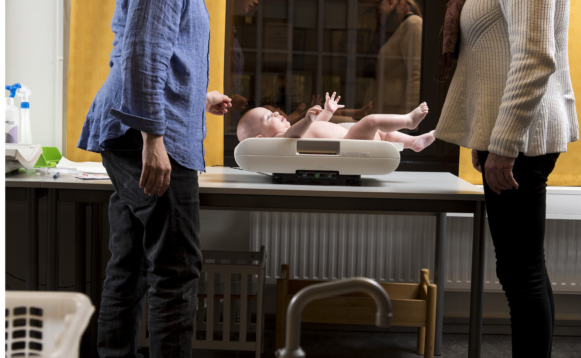 Bildet viser en mor som er på helsestasjonen, der babyen veies av helsesøster.