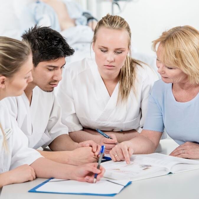 Fasilitator og tre studenter sitter tett sammen rundt et bord. To av dem holder i en penn, og  det er papirark på bordet.