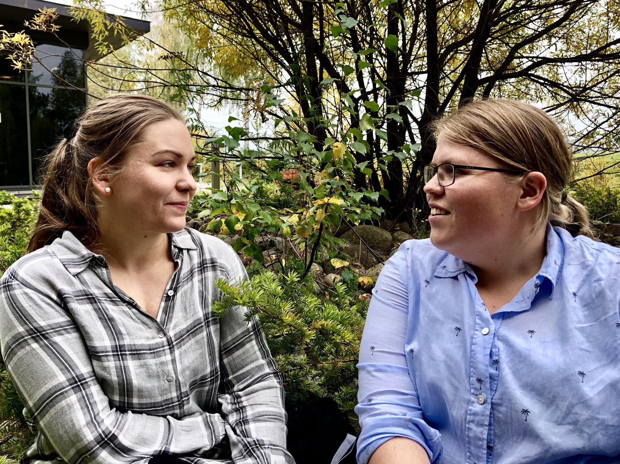 To unge damer se mot hverandre, sitter ute i høstlig landskap  