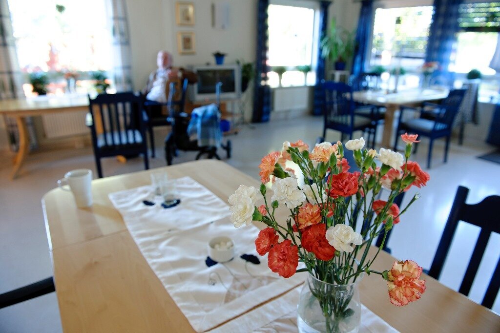 Bildet viser spisesalen på et sykehjem med blomster på bordet og en mannlig beboer i bakgrunnen