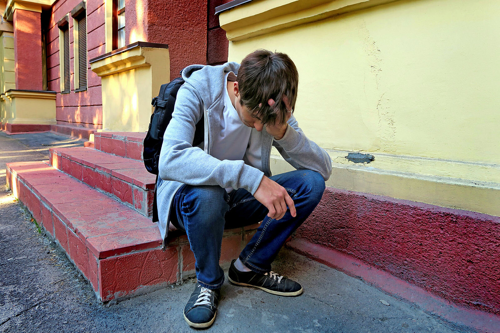 Bildet viser en tenåringsgutt med ransel som sitter på en trapp utenfor skolen. Han er lei seg.