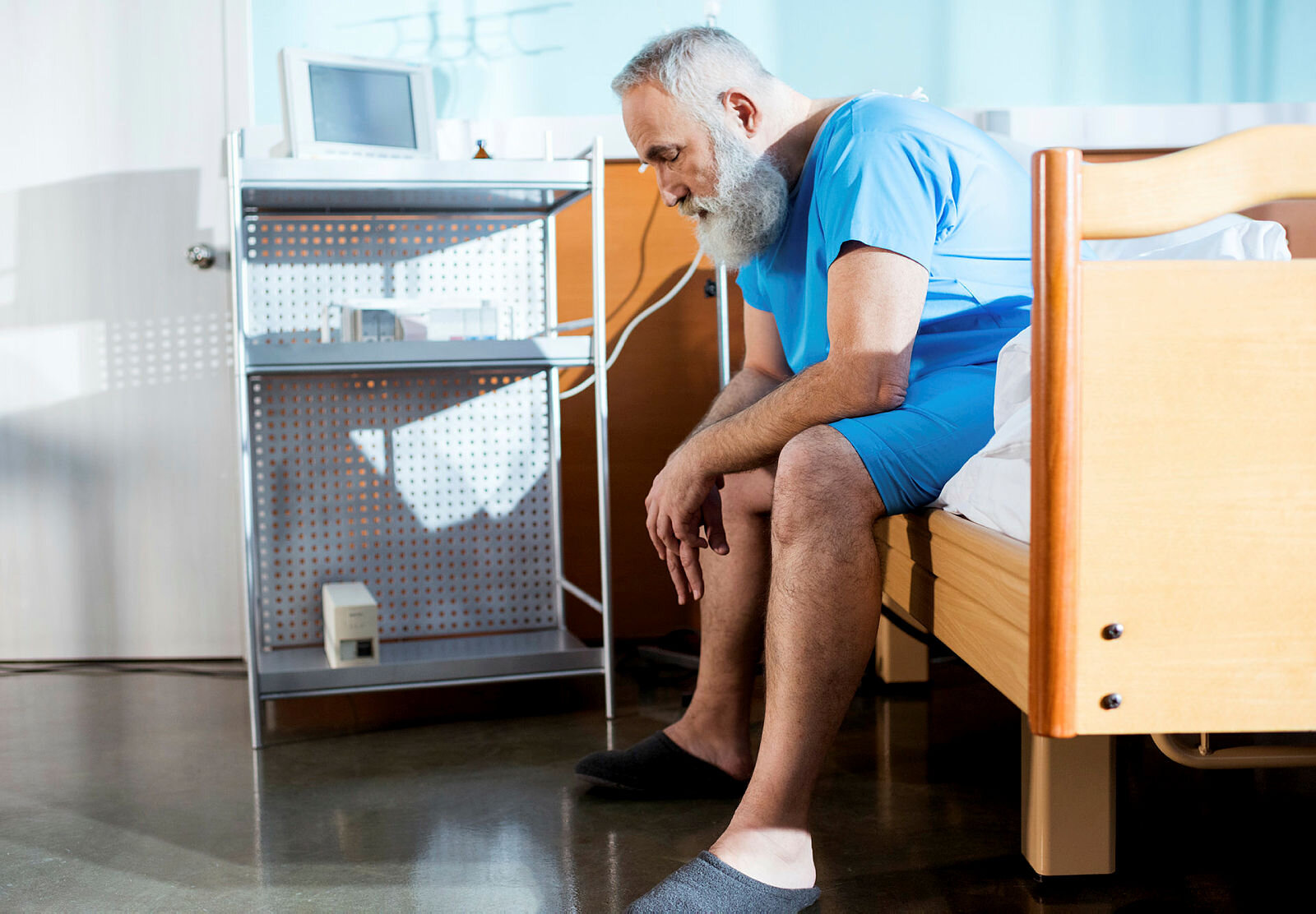 Bildet viser en eldre mann med skjegg som sitter på kanten av en sykehusseng med hendene i fanget.