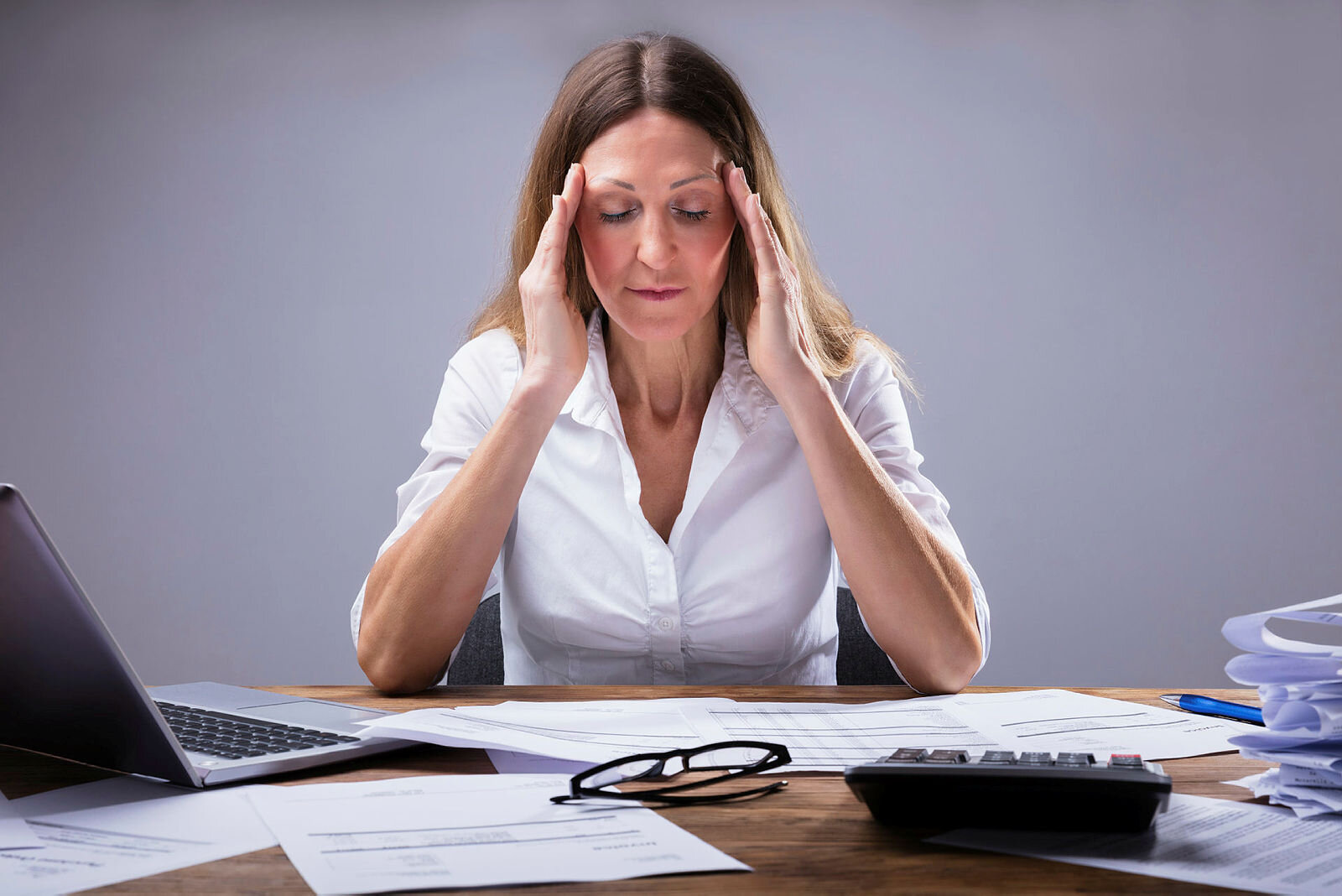 Bildet viser en kvinne som sitter foran et skrivebord med forskjellige permer og rekvisita på og holder seg til tinningene