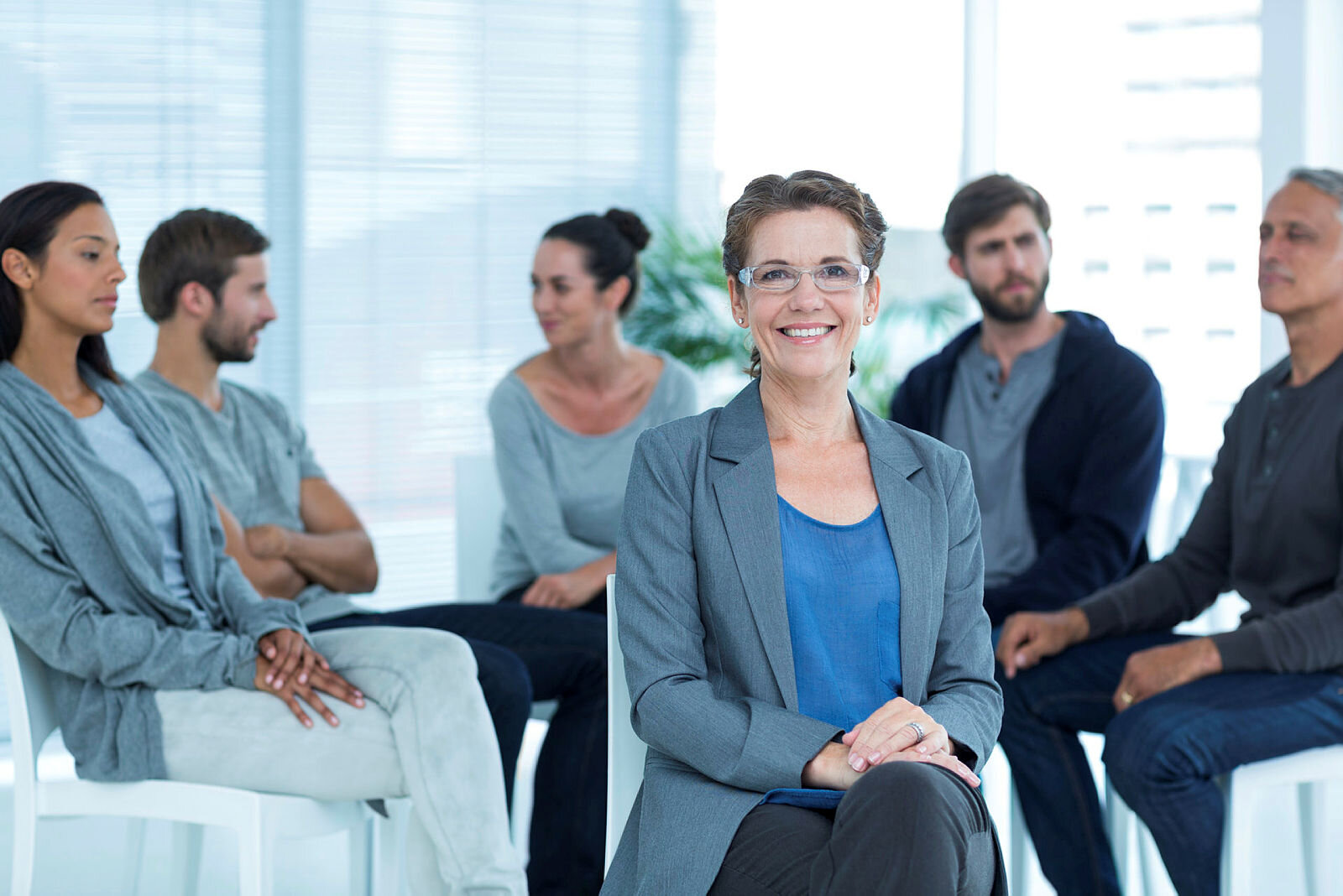 Bildet viser en gruppe som sitter i en halvsirkel og en kvinne som sitter med ryggen til gruppen og ser på oss