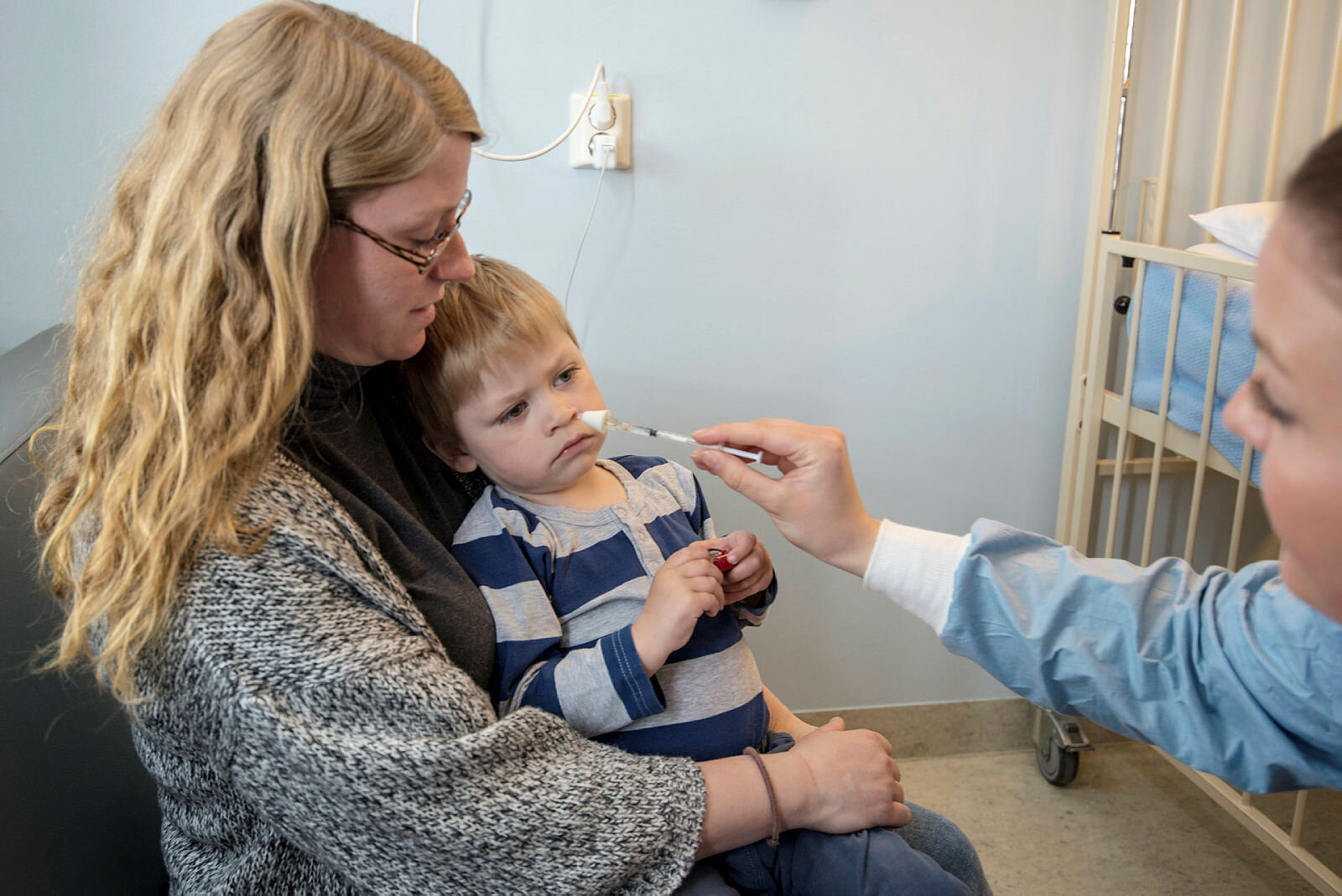 Bildet viser Oliver som sitter på mammas fang og får nesespray av en sykepleier.