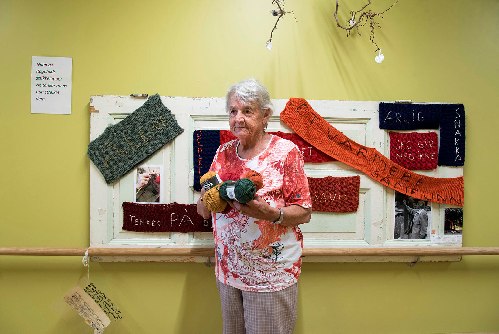 Bildet viser demenssyke Ragnhild på Fosshagen dagsenter i Lier. Hun står foran en tavle med lapper hun har strikket. På lappene står blant annet "alene", "savn" og "ærlig".
