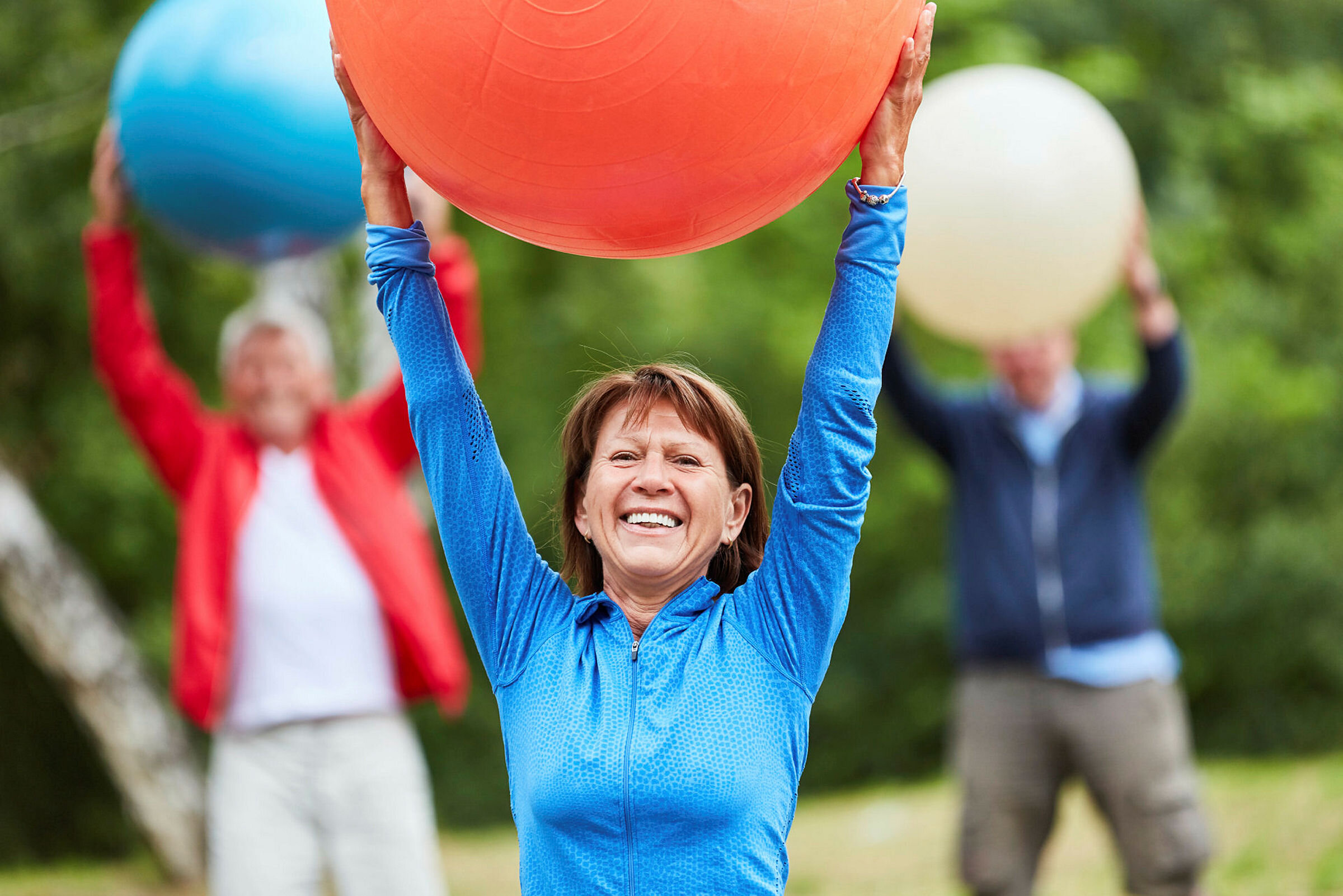 Bildet viser en smilende eldre kvinne i treningstøy som holder en stor ball over hodet.