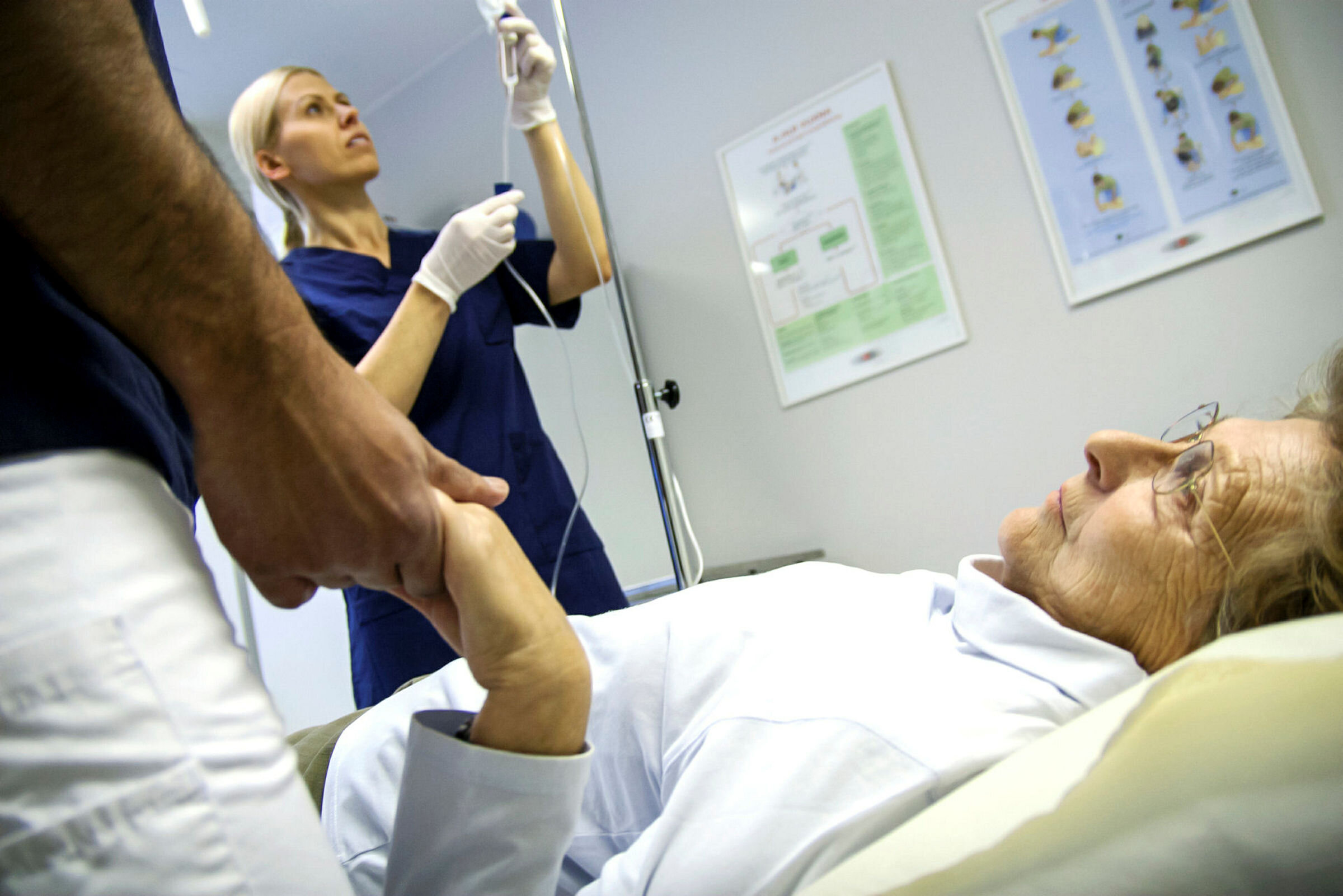 Bildet viser en syk pasient i sengen. Ved sengen står en sykepleieveileder og en sykepleierstudent.