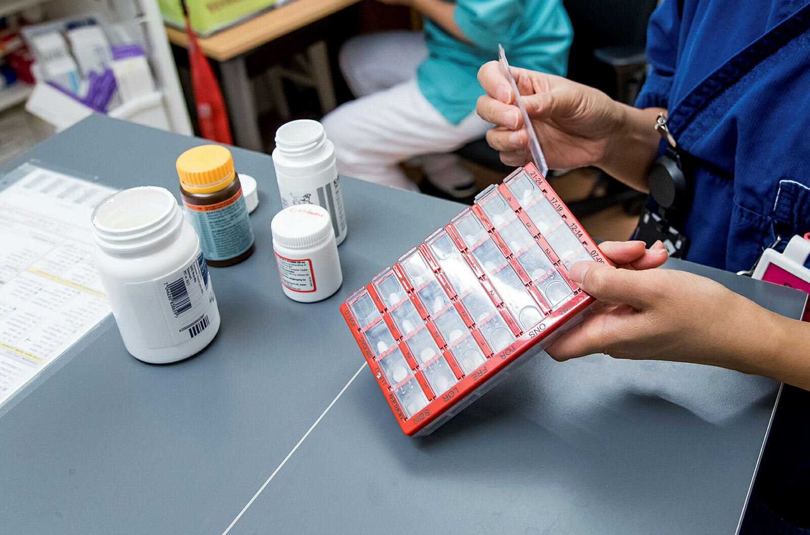 Bildet viser en apoteker/hjemmesykepleier/sykepleier som legger medisiner oppi en dosett. Ved siden av står flere pilleesker.