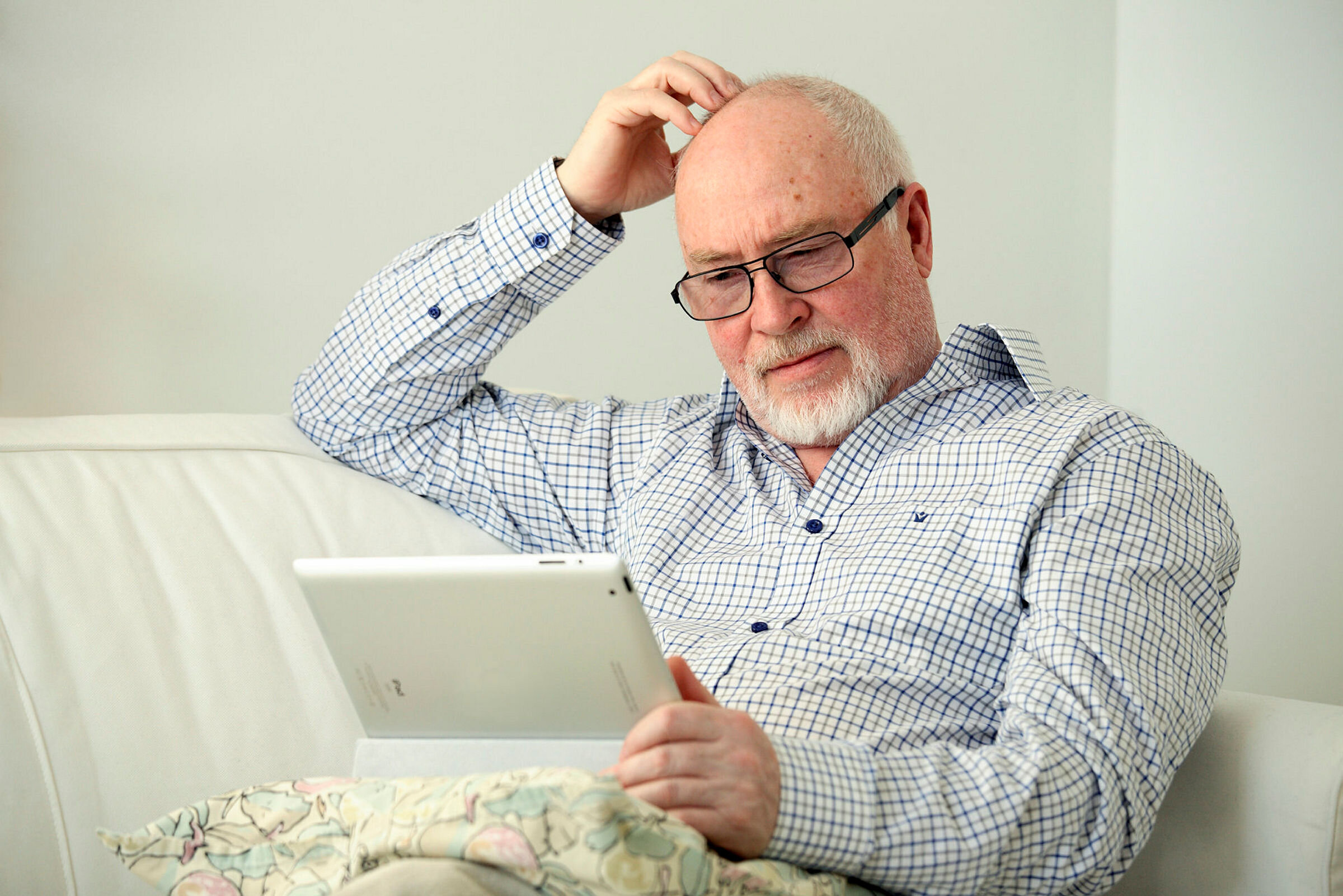 Bildet viser en eldre mann som sitter i en sofa med et nettbrett på fanget og klør seg litt i hodet
