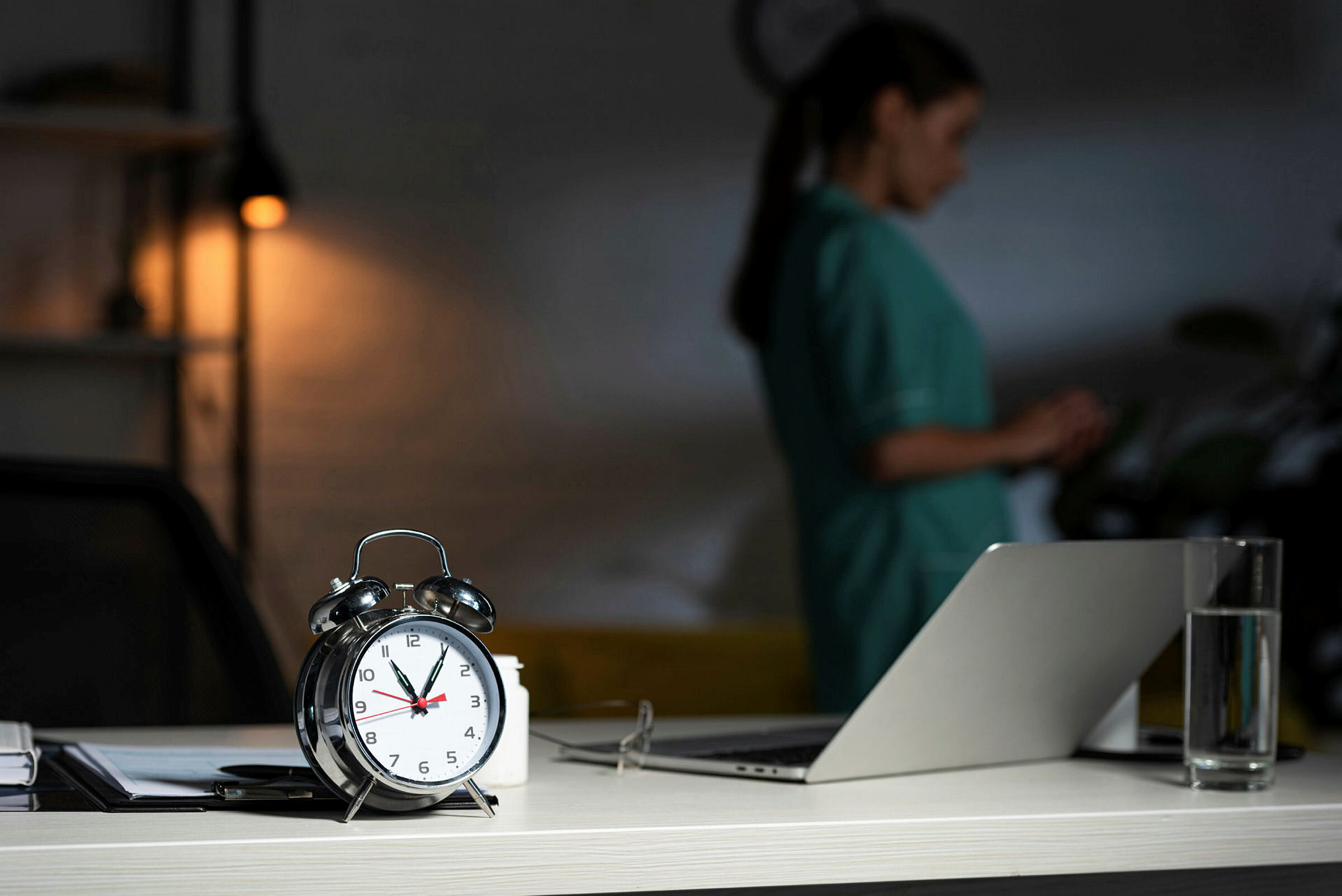 Bildet viser en vekkerklokke i forgrunnen og en sykepleier og en tent lampe i bakgrunnen. Rommet er ganske mørkt.