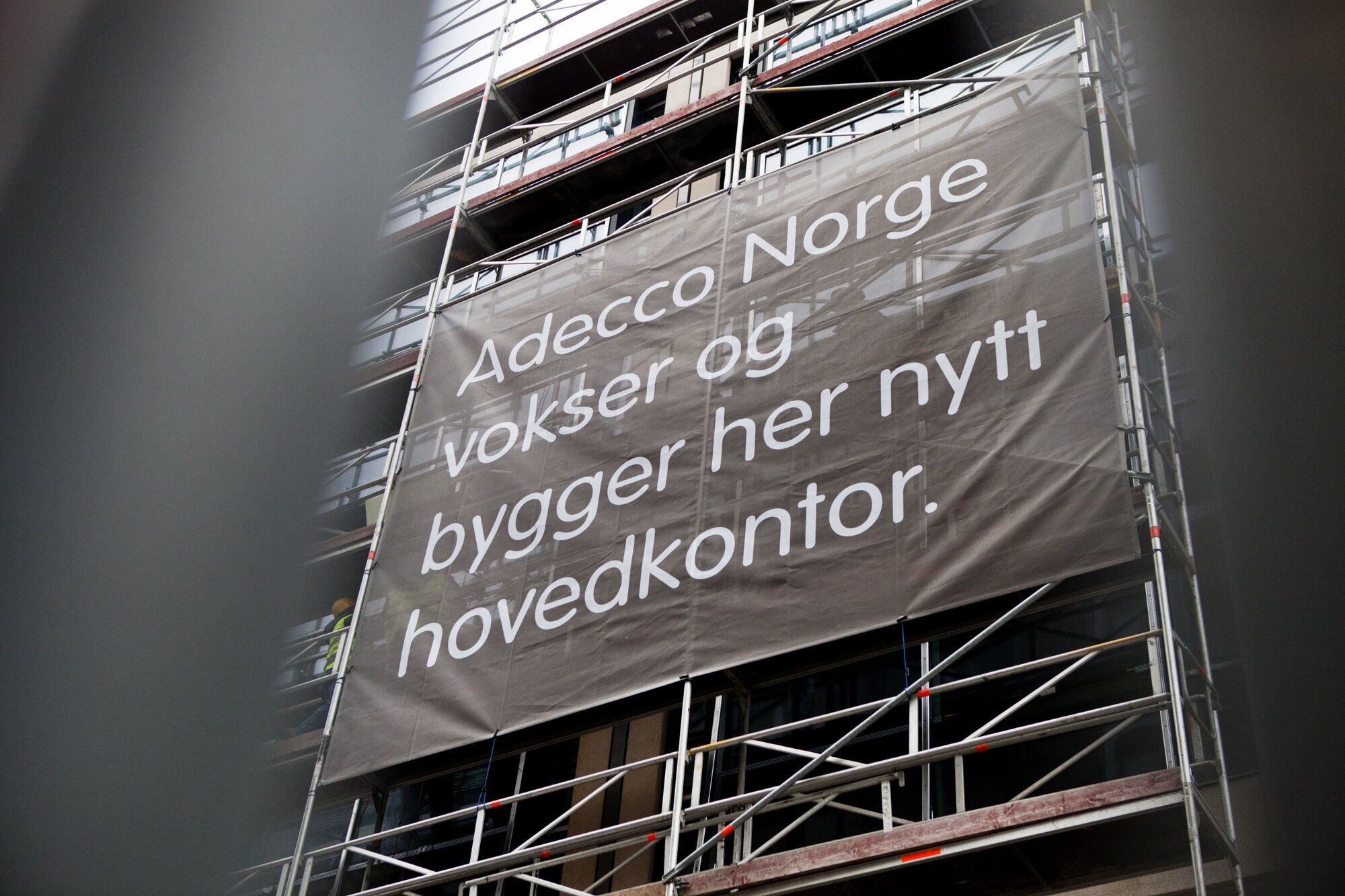 Bildet viser fasaden av et bygg under oppføring, med et banner der det står Adecco Norge vokser og bygger her nytt hovedkontor