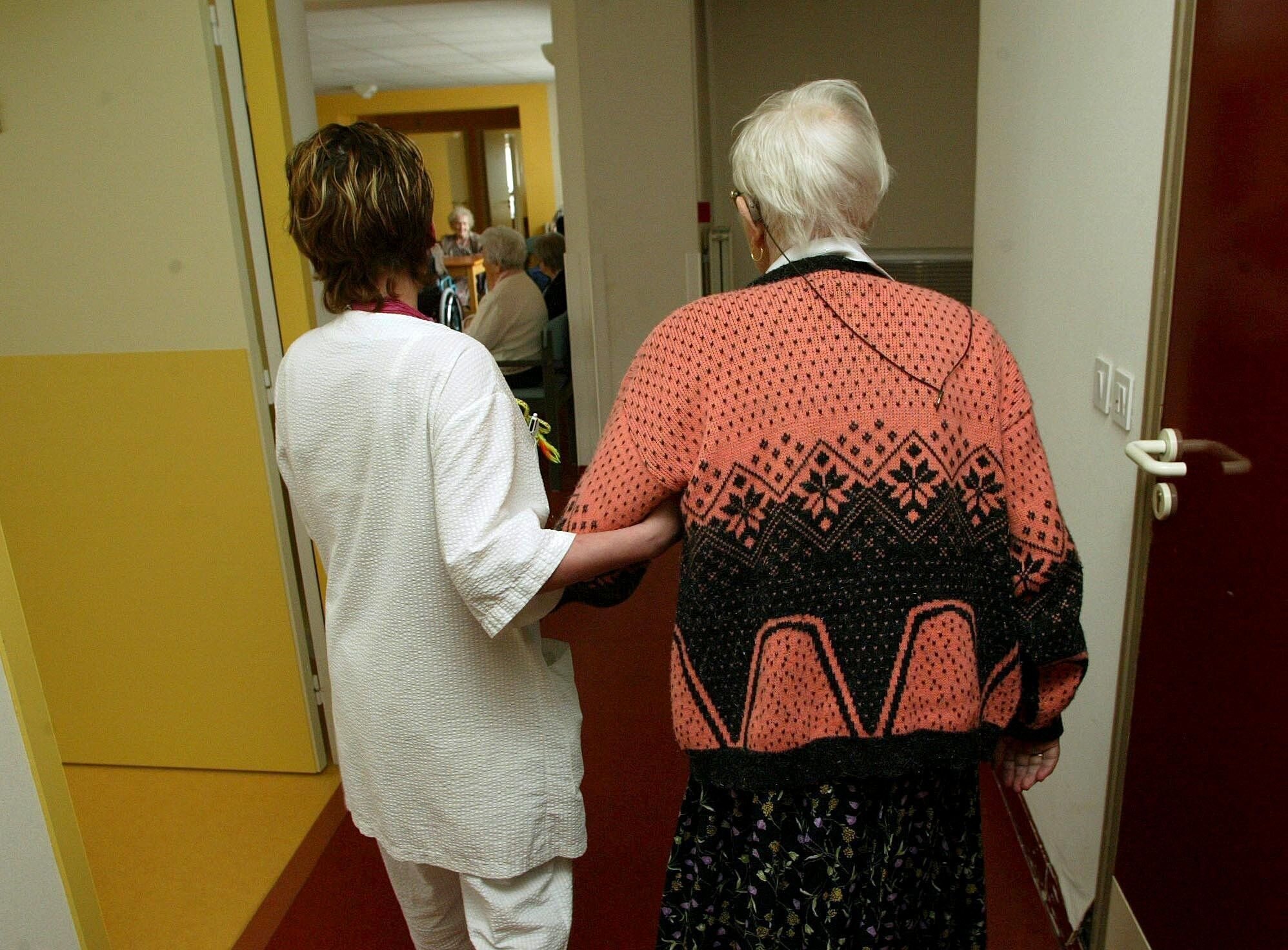 En pleier leier en sykehjemspasient mot spisestuen. Bildet er tatt bakfra.